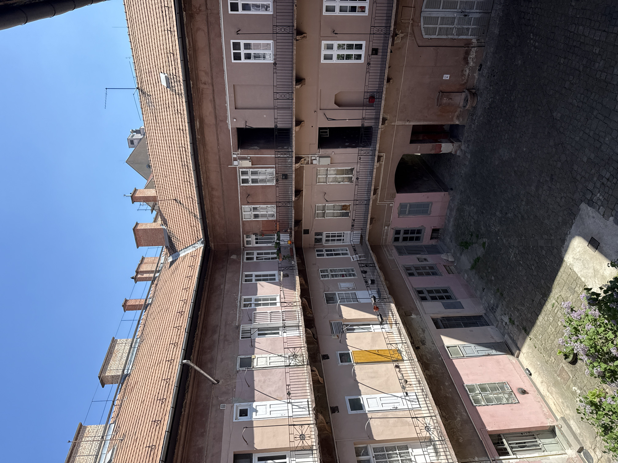 This image captures an overhead view looking down into an inner courtyard of multi-story buildings, typical of urban architecture in Budapest, Hungary. The clear, bright blue sky indicates a sunny day, with strong shadows cast by the buildings across the courtyard below, suggesting it is midday or early afternoon.
The courtyard is enclosed by buildings featuring facades primarily in shades of pale pink and light brown/beige. Multiple levels of these buildings are visible, each with numerous windows, many framed in white, some appearing to have internal shutters. Black wrought iron balconies with decorative patterns extend along many of the building fronts, occasionally showing small potted plants or items.
On the left side of the frame, a prominent terracotta-tiled roof slopes downwards, dotted with several brick chimneys and a television antenna or satellite dish. Beyond this roof, a more distant building structure is partially visible against the sky.
The ground level of the courtyard is paved with dark, uneven cobblestones. A patch of green bushes with small purple flowers, possibly lilac, grows in one corner. In another section of the courtyard, an old-fashioned circular well or water feature can be seen near an arched passageway that suggests an exit or connection to another part of the complex. A small red object, possibly a fire extinguisher, is visible on an interior wall.
No people are present, and no discernible text is visible. The scene conveys a quiet, sunlit urban environment.