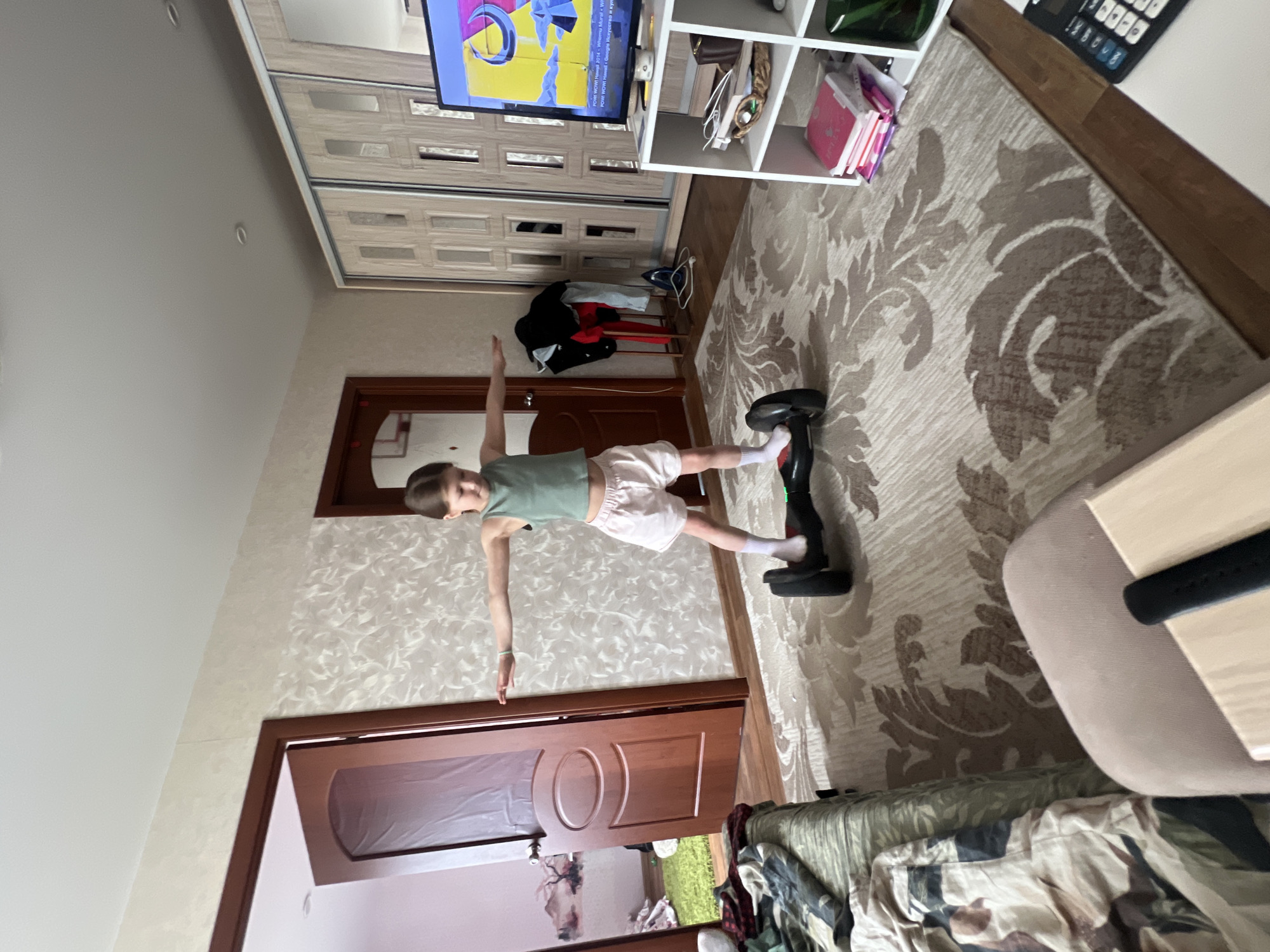 The image, taken in Chaikovsky, Russia, shows a child balancing on a hoverboard indoors, likely in a living room or bedroom. The child, wearing a light green tank top and light pink shorts, has their arms outstretched for stability, demonstrating focus and balance during play or exercise. The black hoverboard has subtle green accents.
The setting features patterned wallpaper and a large patterned rug with a cream background and brown damask-like motifs. In the background, a light-colored wardrobe with sliding doors is visible. Above the wardrobe, a flat-screen television displays colorful graphics and text, which is partially legible as "POW WOW! Hawaii 2014 - Whernu Mural W" and "POW! WOW! Hawaii Google Искусство и кул" (Google Arts & Culture). To the right of the TV, a white open shelving unit holds various items like books and bags.
Further into the room, a clothing rack with clothes, an ironing board with an iron, and two brown wooden doors are visible. One doorway reveals a glimpse of an outdoor area or another room. In the foreground, part of a couch or bed with a camouflage-patterned blanket is visible. The scene is well-lit, suggesting daytime. The overall impression is a casual, lived-in home environment.