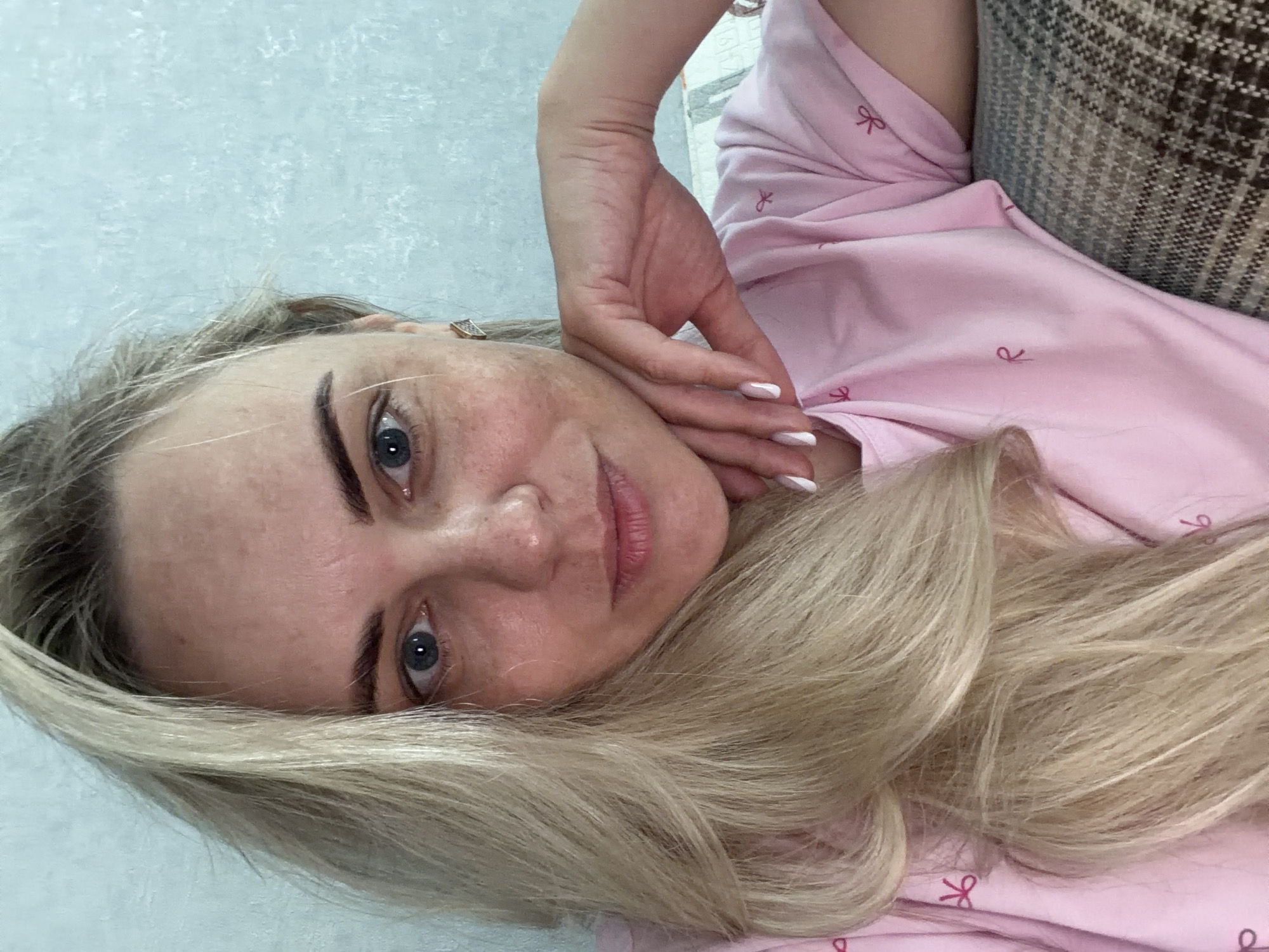 A woman with blonde hair and blue eyes is resting on a surface, wearing a pink top with bow patterns. Her hand rests on her face. The background appears to be a wall and a piece of furniture. The location of the scene is not discernible within Votkinsk, Russia.