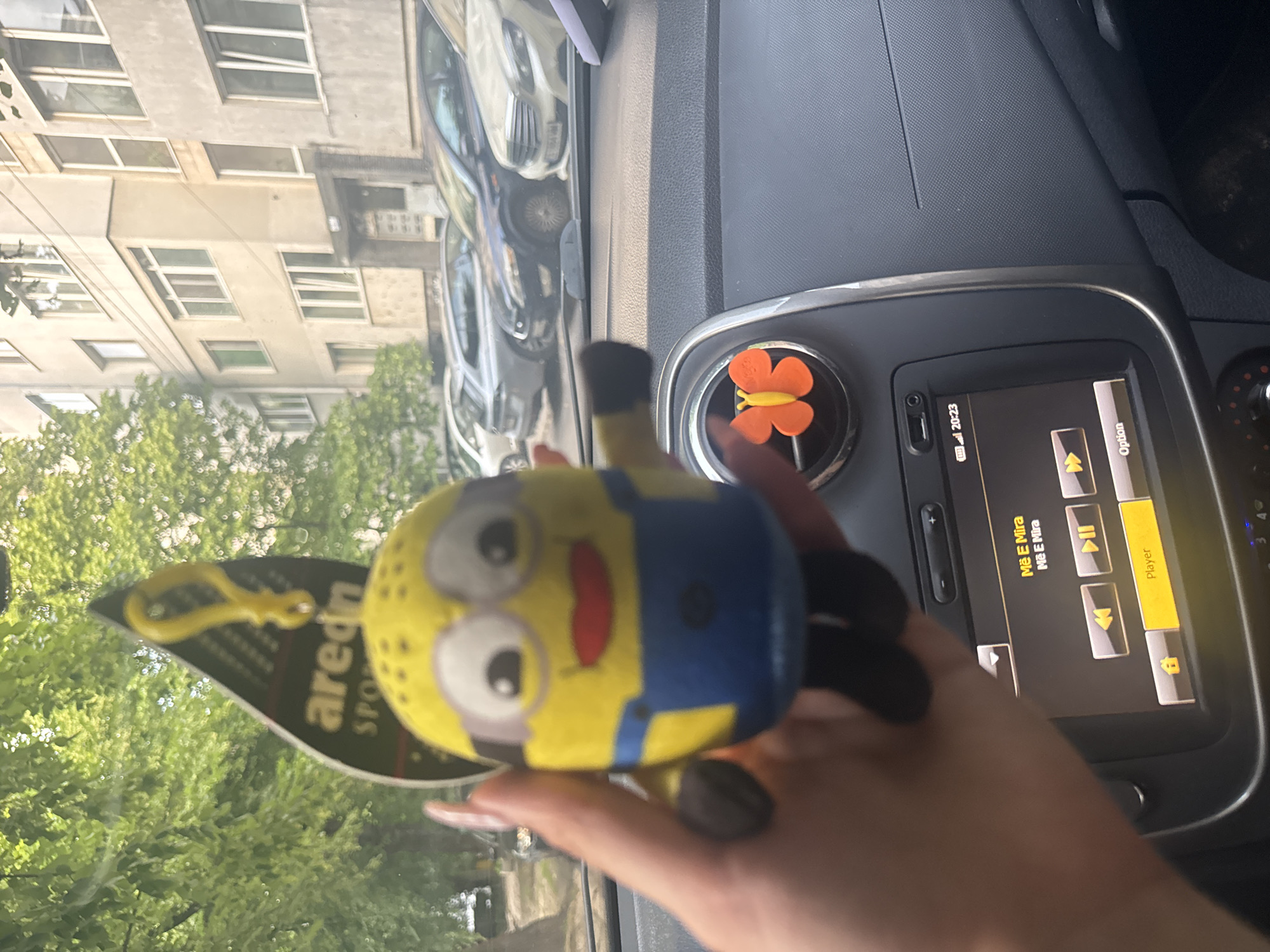 The image, captured from inside a vehicle in Sofia, Bulgaria, depicts a person's hand holding a yellow Minion-shaped air freshener against the backdrop of a residential street. The Minion figure, featuring white eyes, a red smile, and blue overalls, is partially obscuring a black, leaf-shaped AREON SPORT air freshener tag with a yellow plastic clip.
Inside the car, the black textured dashboard is visible, along with an air vent on the passenger side adorned with an orange butterfly clip. The car's infotainment screen, displaying the time 20:23, shows the title "Më E Mira" (possibly a song title) and standard music player controls, including play/pause and skip buttons.
Outside the windshield, a multi-story, light-colored residential apartment building, characteristic of Eastern European architecture, stands across the street. Numerous windows are visible on its facade. Parked parallel to the curb in front of the building are several cars, including a silver Mercedes-Benz with the Bulgarian license plate `CB 7886 MH` and a dark blue Ford model. Lush green trees line the street, suggesting spring or summer conditions. The ambient light indicates late afternoon or early evening.