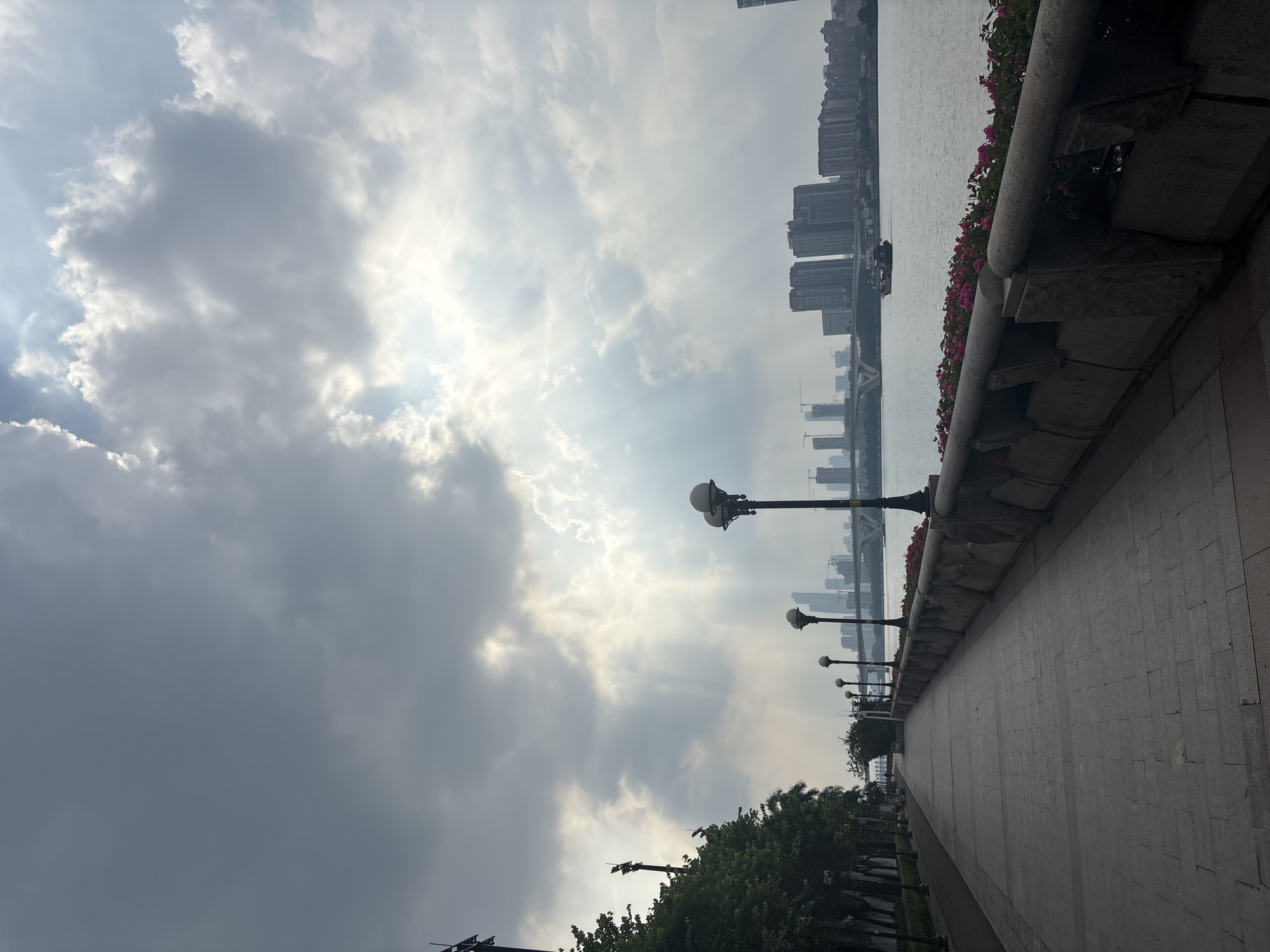 The image depicts a scene along a waterfront promenade, likely in Haizhu District, China. The perspective is from ground level, looking towards a river and a cityscape in the distance. The sky is filled with dramatic, sun-dappled clouds, suggesting a day with intermittent sunlight, possibly late afternoon or early morning.
The foreground features a paved walkway with a decorative stone railing on its left. Pink flowers are planted along the railing, adding a splash of color to the scene. Several ornate streetlights with multiple globes are spaced along the promenade, hinting at its use for evening strolls. To the left of the walkway, there are trees and more streetlights, suggesting a park-like or landscaped area.
Across the river, a dense collection of modern high-rise buildings forms the city skyline. Some of these buildings appear to be residential or office towers, characteristic of a major urban center. The water itself is calm, reflecting the cloudy sky.
There are no people visible in the image. The overall mood is serene and slightly overcast, with the natural beauty of the sky and the man-made structures of the city coexisting. The time of day is suggested by the quality of light breaking through the clouds, casting rays that highlight the atmospheric conditions.