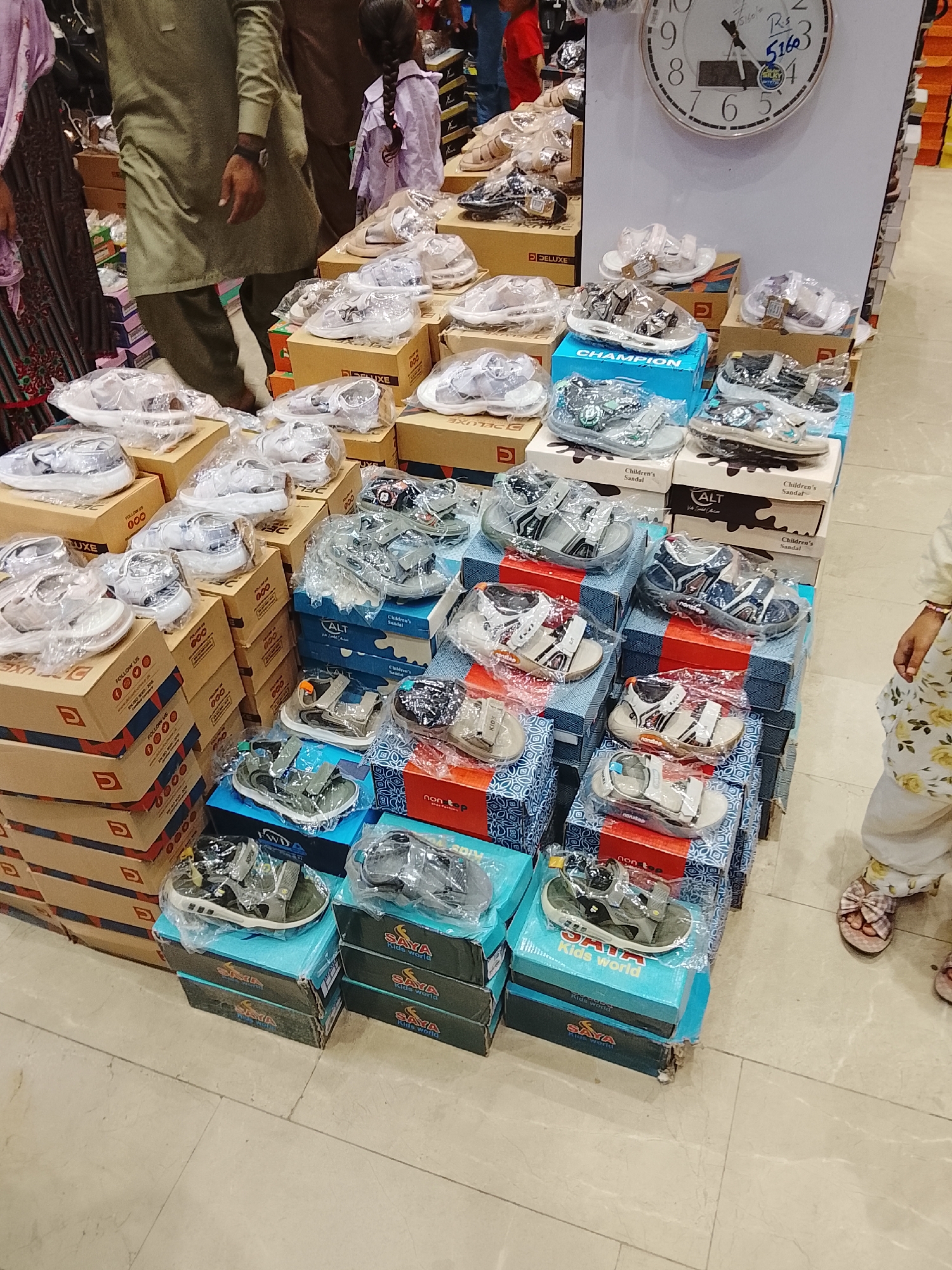 The image shows a collection of children's sandals displayed in a retail setting, likely a shoe store in Gulgasht Colony, Pakistan. Numerous boxes, stacked to varying heights, hold the sandals. The boxes feature logos and brand names, including "LUXE," "DELUXE," "CHAMPION," "ALT," "non stop" and "SAYA Kids world." Many of the sandals are individually wrapped in clear plastic.
Several individuals appear in the background, including a person wearing a light green shalwar kameez, a girl with a braided ponytail, and other partially visible figures.
A large round clock hangs on a white wall above the shoe display, displaying the time around 11:00 and a price tag of "Rs. 51603."
The floor is tiled in a light color. A child with a floral-patterned dress and sandals stands near the display.