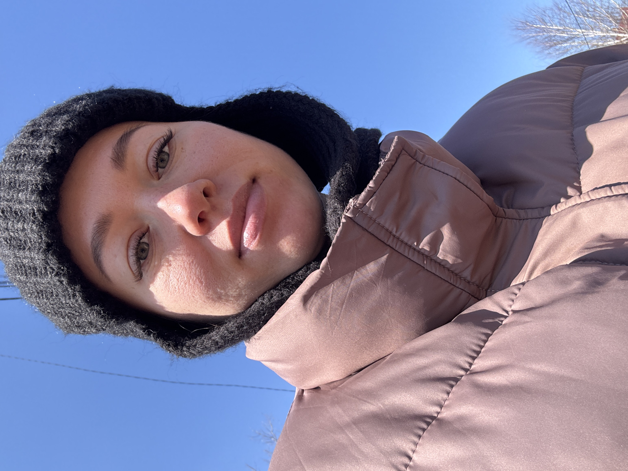A low-angle, close-up selfie shows a person with light skin, wearing a black knit hat and a puffy, brown jacket against a backdrop of a clear, blue sky. Bare tree branches are visible in the upper right corner. No specific details indicate that the location is in the city of Ferma, Russia.