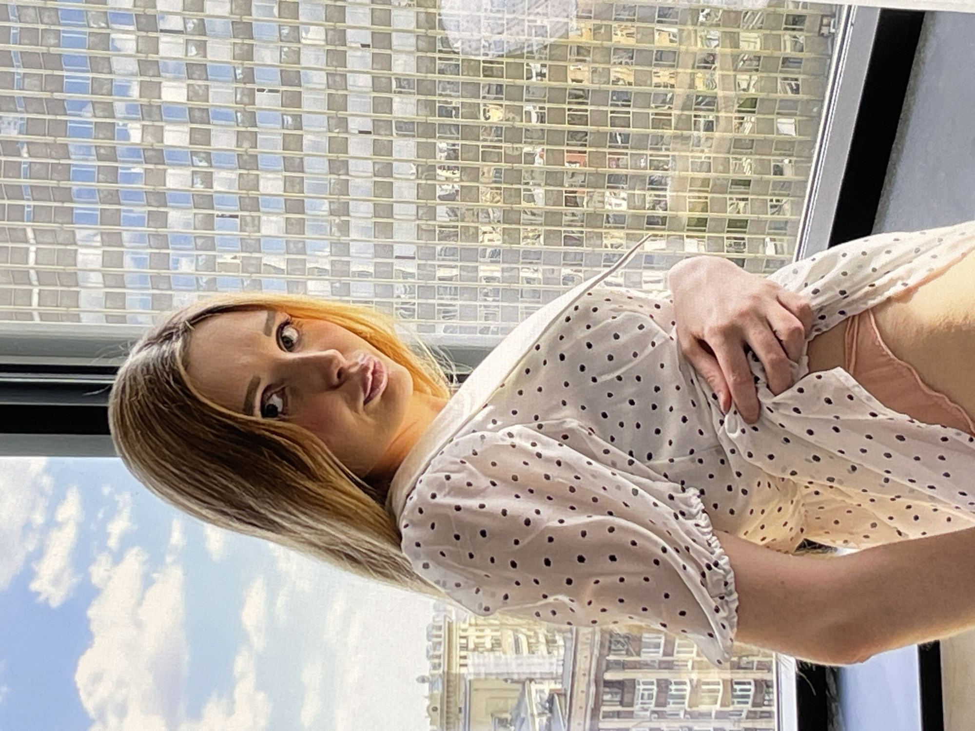 In an urban setting, a blonde woman stands near a large window. She is wearing a white dress with black polka dots and has her hand on the hem of the dress, lifting it slightly to reveal her peach-colored underwear. The window overlooks a cityscape with buildings and a sky filled with clouds. The woman is looking directly at the camera with a light makeup application.