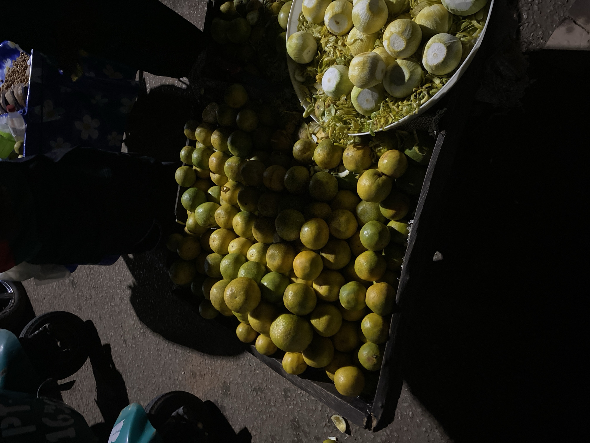 Captured at night in Bukuru, Nigeria, the image shows a mobile street vending setup. A metal cart is heavily loaded with two types of citrus fruits. On the left side, there is a bulk of whole oranges piled high. To the right, a white bowl overflows with peeled oranges, surrounded by the discarded citrus peels. The cart sits on a paved surface, presumably a street or sidewalk. A portion of a blue, patterned cloth and a container of small, unidentified objects are visible on the left side. Parts of what looks like a turquoise motorbike are also visible. The overall scene is dimly lit.
