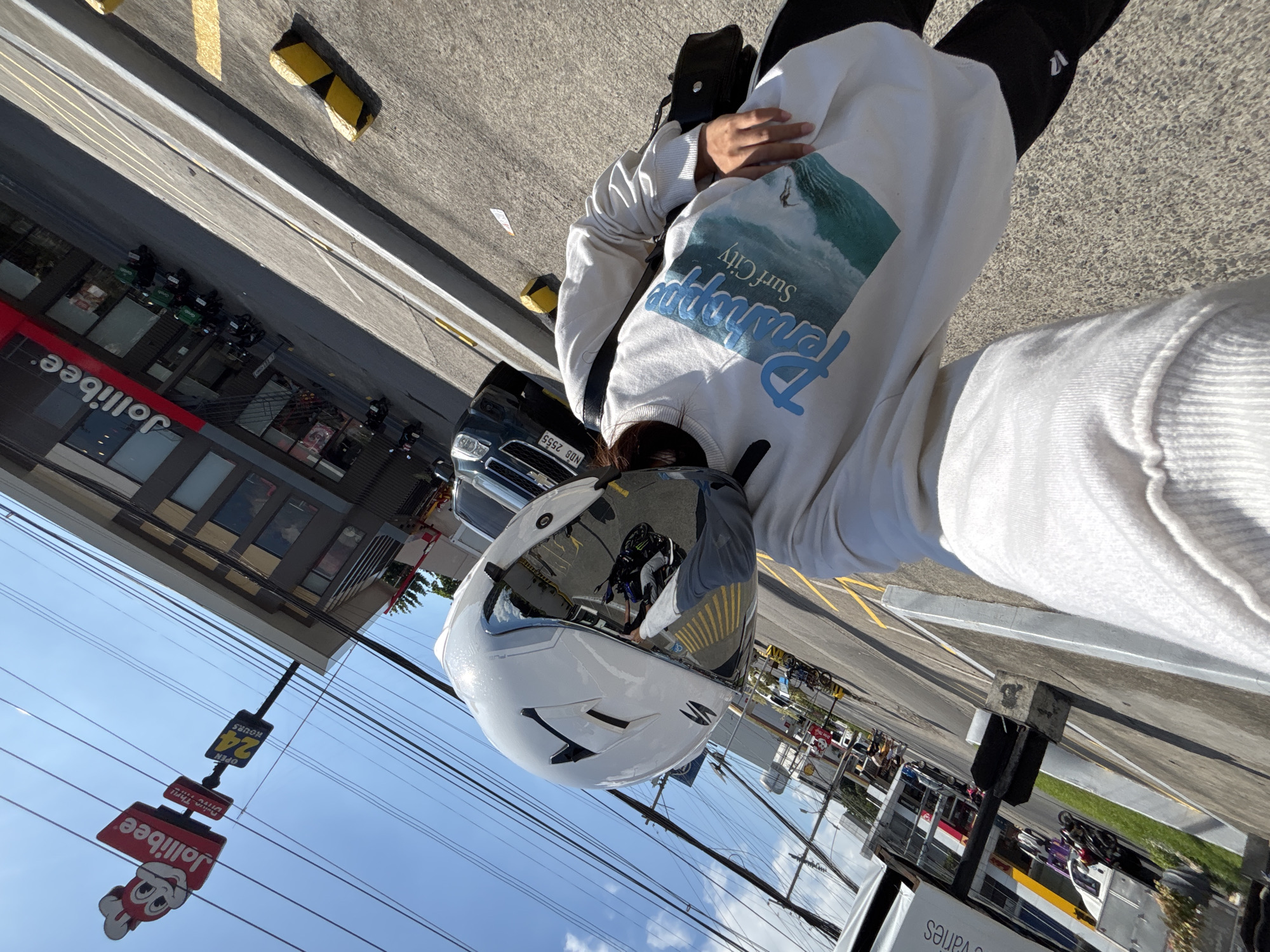 A person wearing a white motorcycle helmet with a reflective visor, a white sweatshirt with a graphic of waves and the text "Penang Surf City", and black pants is taking a selfie outdoors. The person is holding a black bag with a strap over their shoulder. The background shows a Jollibee restaurant and a street with parked motorcycles and a car. The scene is likely in Davao City, Philippines, indicated by the Jollibee branding and the presence of motorcycles commonly used for transportation in the Philippines. The environment appears to be a bright, sunny day.
