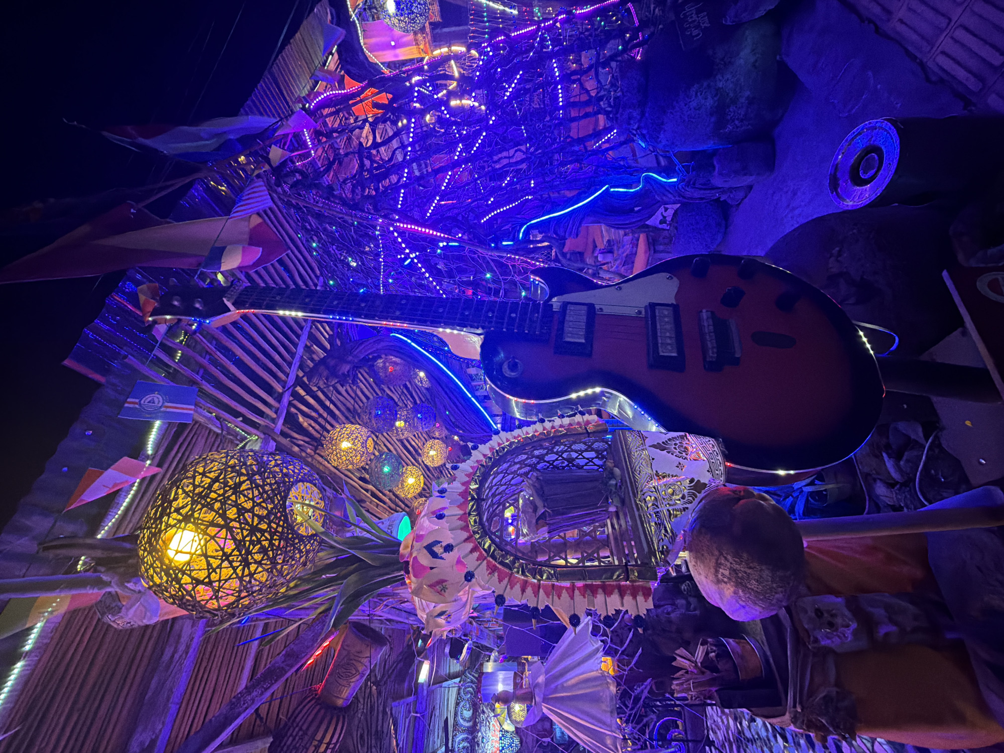 The image captures a dimly lit, atmospheric scene within an establishment in Banjar, Indonesia, likely a bar or a unique lounge. The dominant colors are deep blues and purples from extensive neon and string lighting, accentuating a bohemian and somewhat mystical ambiance. A striking red electric guitar stands prominently in the foreground, angled slightly to the right, with its body and neck clearly visible. Behind it, a complex, vine-like structure is heavily adorned with numerous small, colorful LED lights.
The background reveals a rustic, thatched roof structure, with exposed wooden beams and corrugated metal sheeting. Several spherical, woven lamps with warm yellow light hang from the ceiling, casting a soft glow. Small flags, appearing to be international or symbolic, are strung between supports. To the left, a decorative, fan-like structure with pink and white accents frames a shadowy interior space. Further left, a white, draped object resembling a lampshade or a piece of decor is visible. In the lower left, a round, textured object, possibly a decorative orb or a fruit, is mounted on a wooden stick, resting on an orange cloth. The floor appears to be a tiled or stone surface. The overall impression is one of a vibrant, eclectic, and possibly artistic space designed for entertainment or socializing.