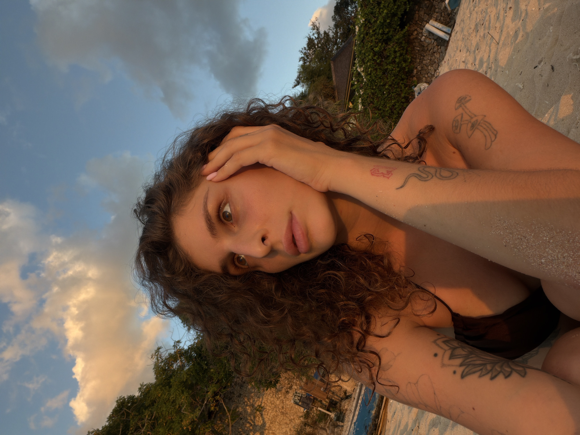 A young woman with curly brown hair and tattoos on her arm is sitting on a sandy beach, her hand resting on her face. She is wearing a dark swimsuit. The sky above is a mix of blue and orange clouds, suggesting it is either sunrise or sunset. Lush green trees and a stone wall are visible in the background, along with a building and some blue boats on the sand. The lighting is warm and golden.