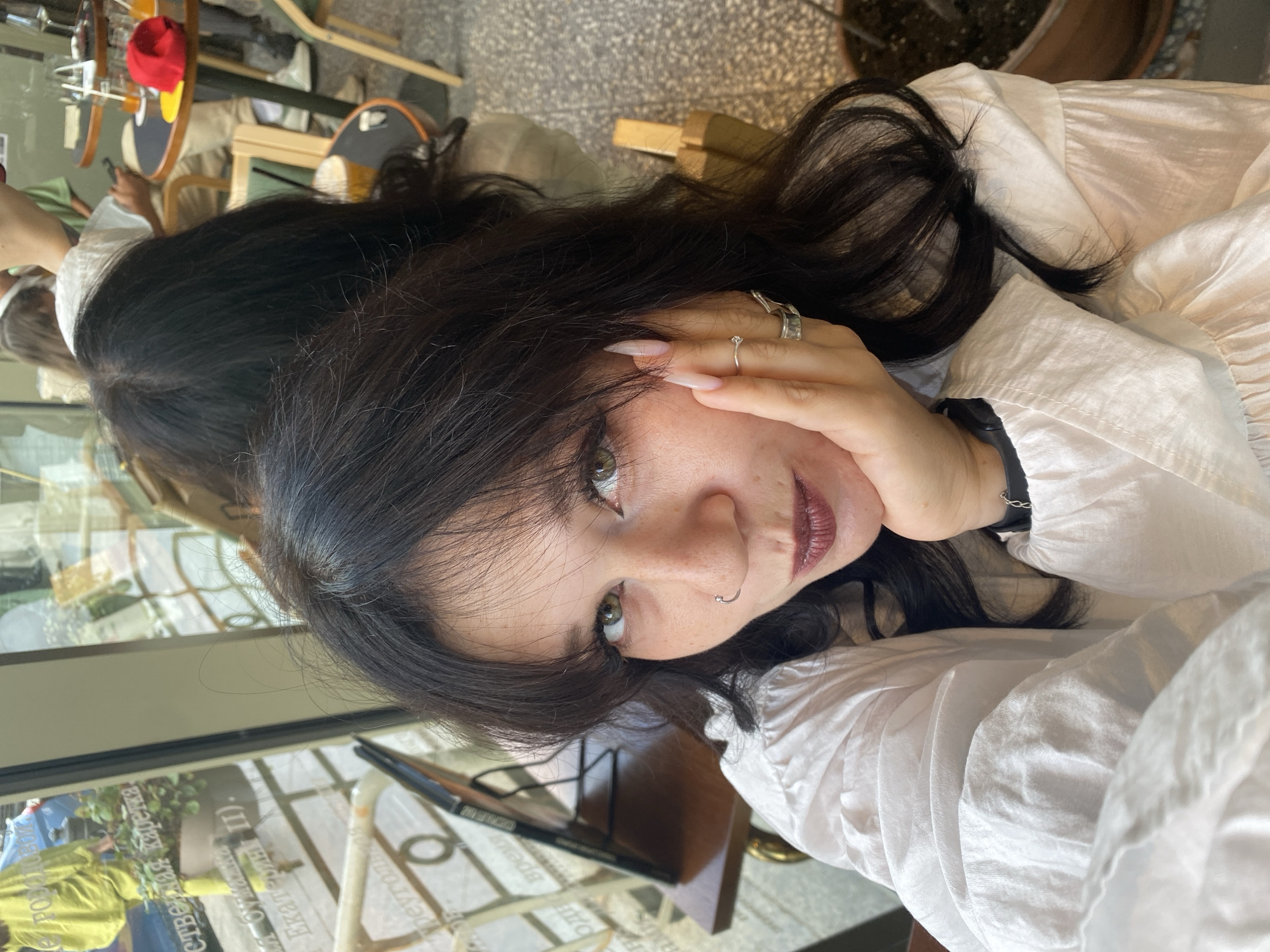 The image is a close-up selfie of a young woman with dark hair and light eyes, wearing a white collared shirt. She is leaning her head on her hand, revealing some rings on her fingers and a nose ring. The background suggests an indoor cafe environment, with tables and chairs visible. Through a window in the background, elements of Saint Petersburg, Russia can be identified. Specifically, text on a poster or windowpane is visible in Cyrillic script, and a blue car is parked outside, common in urban Russian streetscapes. The overall lighting is bright and natural, suggesting daytime.