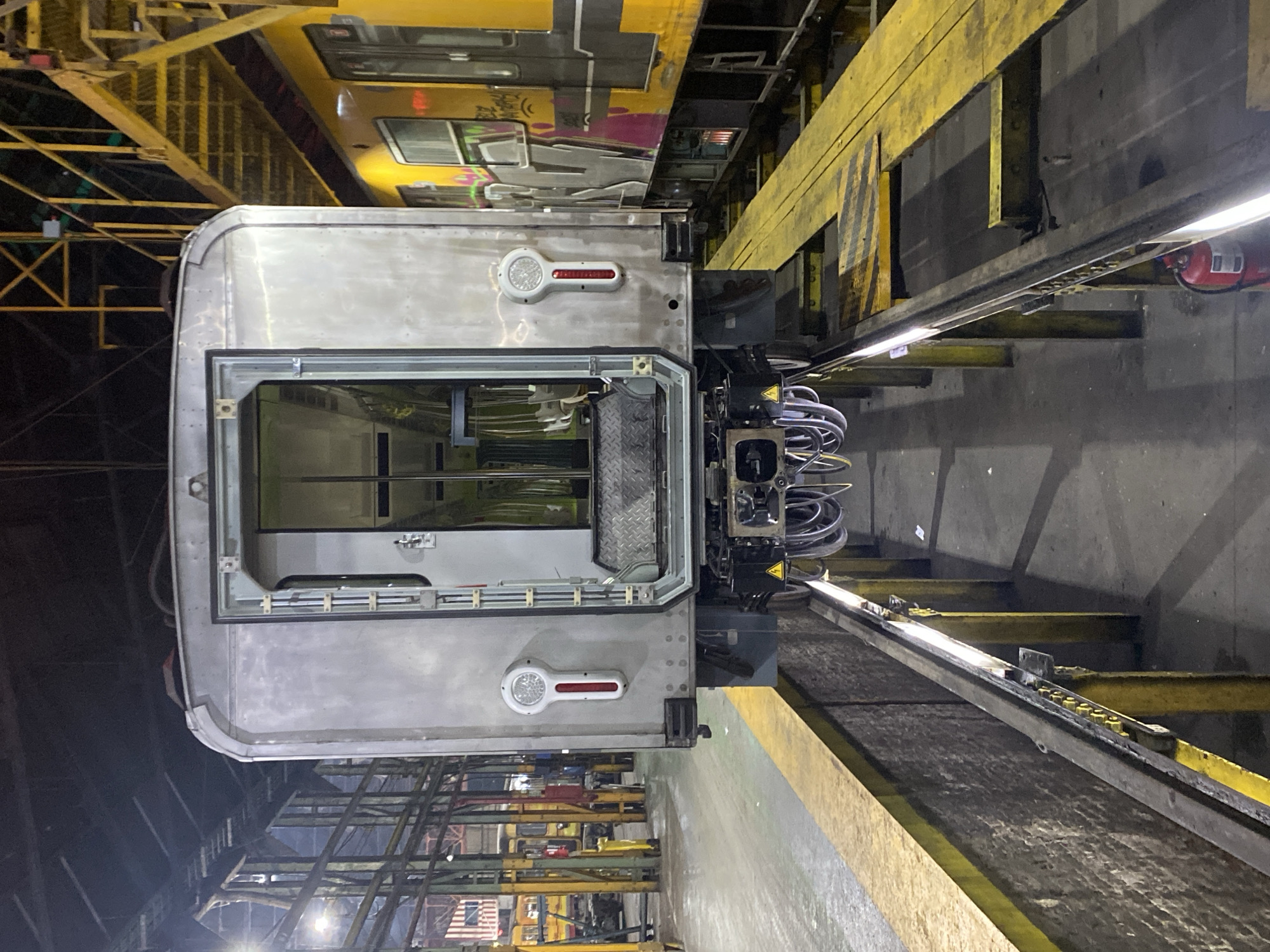 The image shows the rear end of a train carriage, likely undergoing maintenance or repair in a depot. The carriage has a dull silver exterior and features taillights. The rear door is open, revealing the interior. The carriage is connected to a heavy-duty coupling mechanism with multiple cables. The scene is in an industrial setting. There are support beams and scaffolding painted yellow, as well as railway tracks. Another train car can be seen in the upper part of the image. Without further information it is difficult to pinpoint the specific location within Buenos Aires, but it is likely a railway maintenance facility or depot.