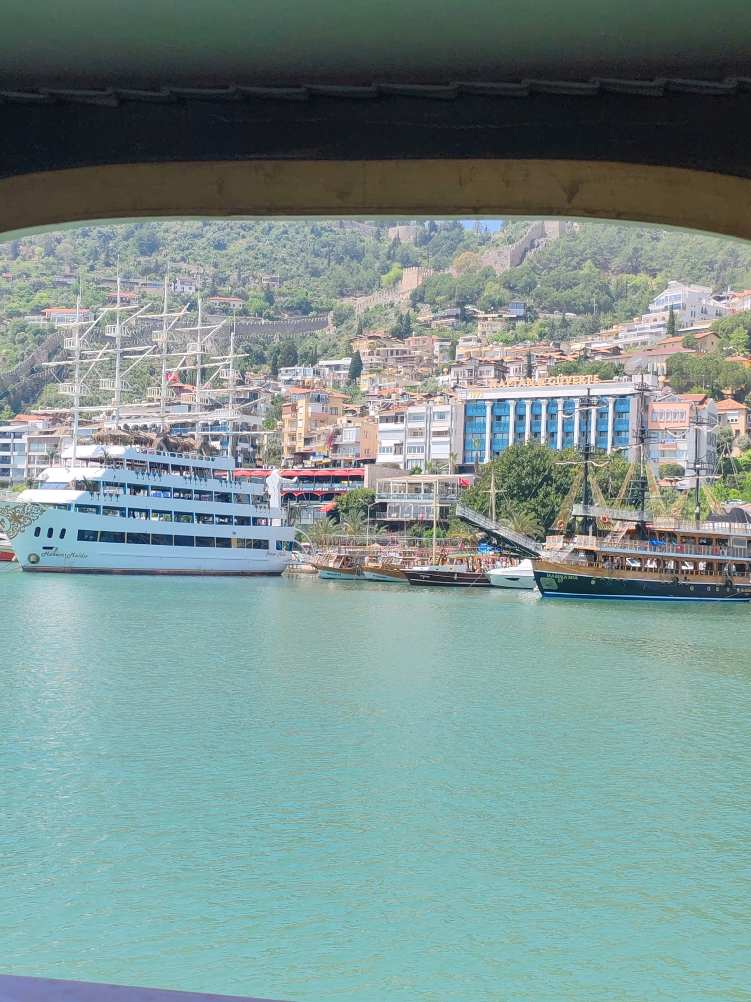 The image shows a harbor scene in Alanya, Turkey, viewed through an opening. The water is a turquoise color and dominates the lower portion of the view. In the middle ground, there are three boats docked at the harbor. The boat on the left is a multi-decked, primarily white cruise ship with numerous masts. To the right of this large ship are two smaller vessels, one appearing to be a historical sailing ship design. In the background, there is a densely populated hillside with buildings of varying sizes and colors. Part of a stone fortress wall is visible on the hillside among trees and buildings. The scene is bright and suggests a clear day.