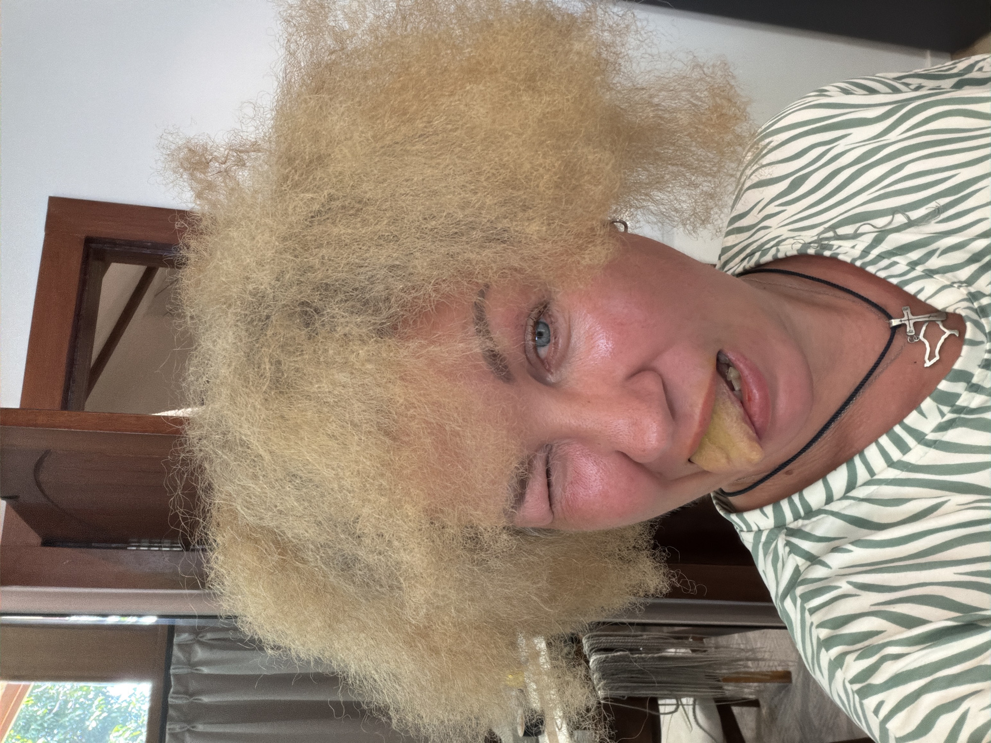 A person with frizzy blonde hair is winking and sticking out their tongue. They are wearing a green and white striped top and a necklace with a silver pendant. The setting appears to be indoors, with a dark wood frame and a window with curtains visible in the background. The location is Unknown, Unknown.