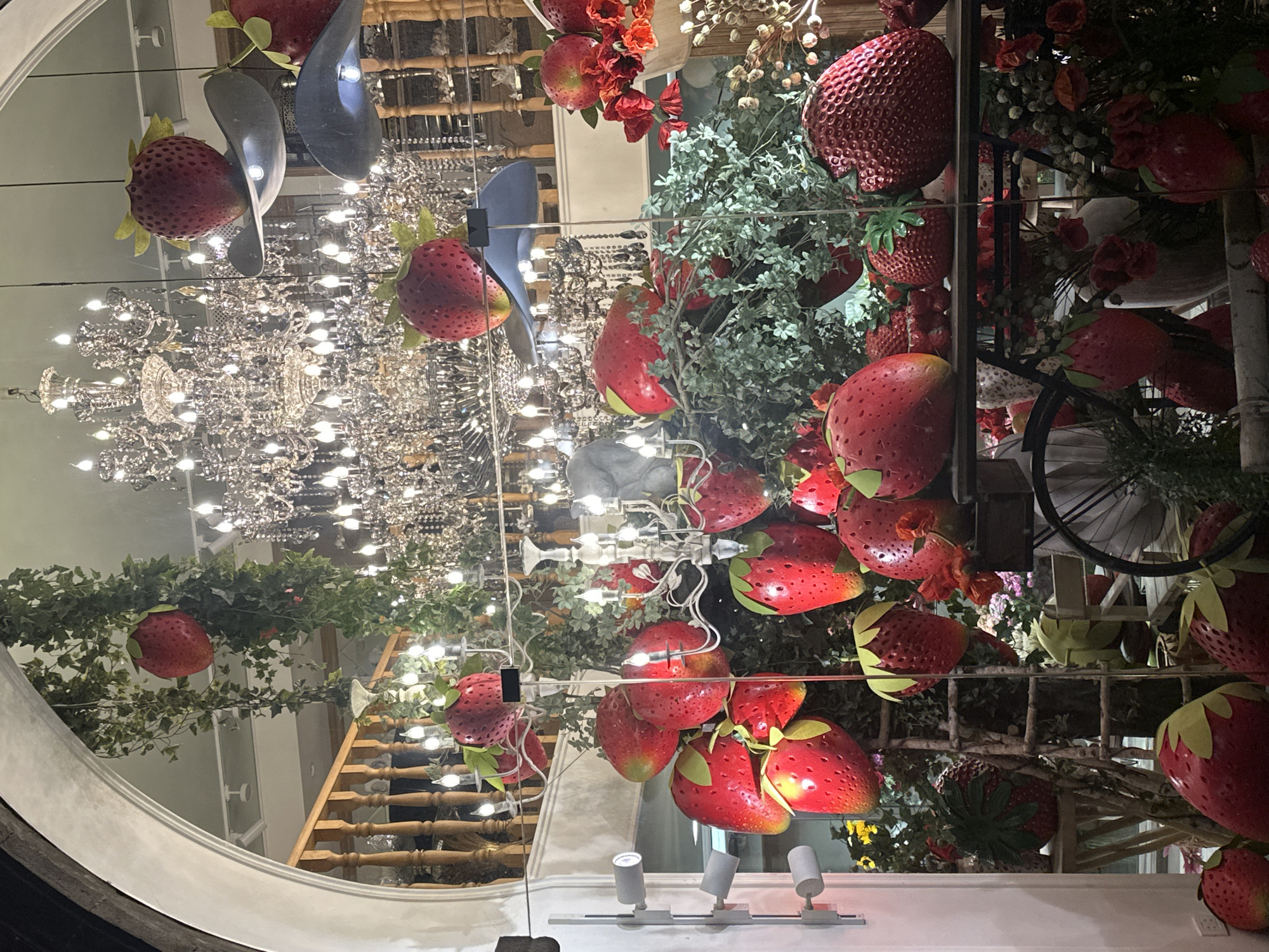 The image captures the reflection of a store window display in Yerevan, Armenia. The display is elaborate, featuring oversized artificial strawberries scattered among greenery, flowers, and decorative elements. A chandelier is prominently reflected, suggesting a sophisticated interior setting. The reflection also reveals details of the store's interior, including a wooden staircase and shelving. The display is illuminated by track lighting, which is visible near the bottom of the frame. The overall impression is one of whimsy and luxury.