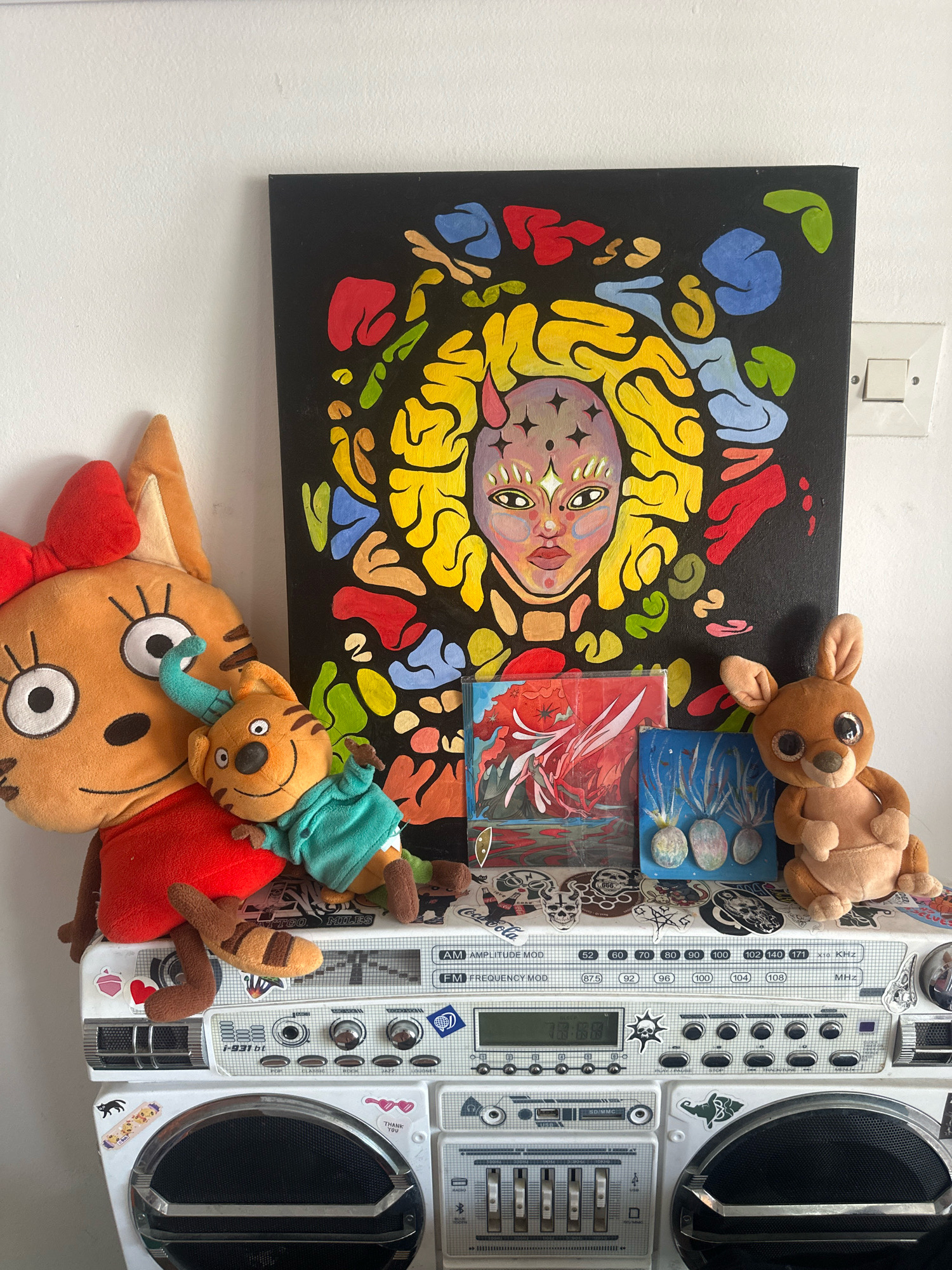 The image displays a white boombox adorned with various stickers. On top of the boombox, there is a canvas painting featuring an abstract design with a central face. To the left of the painting are two plush toys: a large orange cat-like toy wearing a red shirt and a smaller orange tiger-like toy in a teal shirt. To the right of the painting are two small framed artworks and a brown plush kangaroo toy. The boombox and its contents are positioned against a white wall, where a light switch is visible on the right. The scene does not provide any specific indicators of location in Budva, Montenegro, or any actions or interactions occurring.