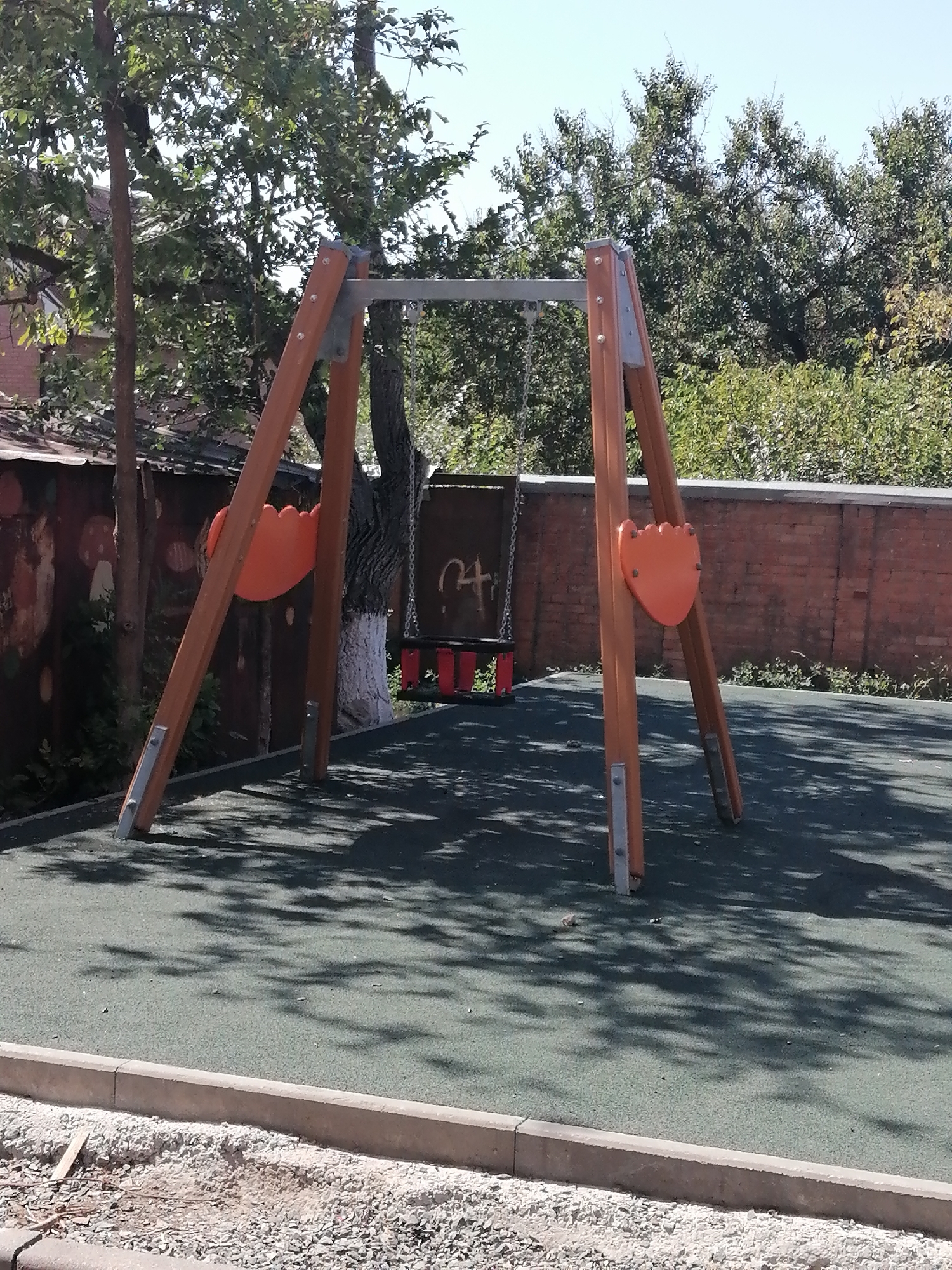 A playground swing set with two orange seats is pictured on a rubberized surface in Rostov-on-Don, Russia. The swing set has wooden A-frame supports connected by a metal bar. A red swing seat hangs from the metal bar. The background features a brick wall, trees, and a chain-link fence. The ground in front of the playground is covered in gravel and some construction materials.