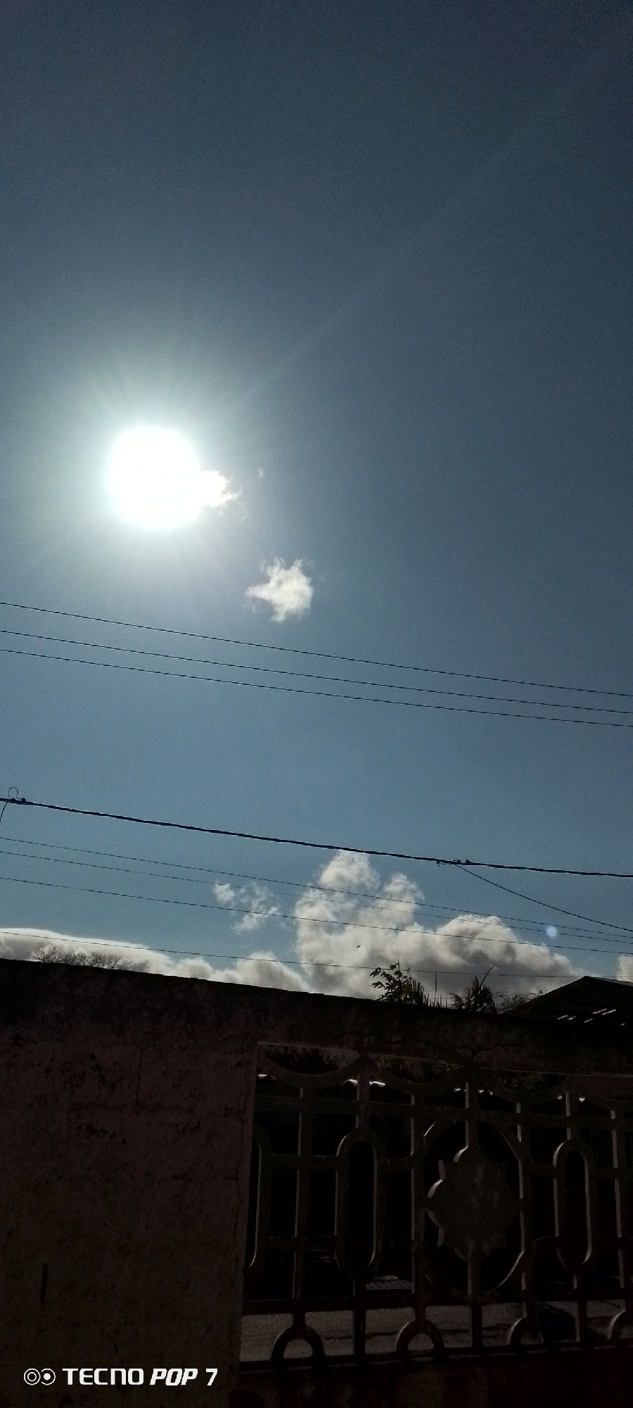 The image presents a bright, sunlit daytime scene, likely in Altagracia, Venezuela, viewed from a low angle, looking up towards the sky and over a residential boundary. The sky dominates the upper portion, featuring a clear, light blue expanse. The sun is a powerful, overexposed white orb in the upper left quadrant, casting bright rays and some lens flare, indicating intense daylight. Scattered across the sky are a few small, fluffy white clouds, one notably close to the sun.
Multiple dark power lines run horizontally across the middle of the frame, traversing the blue sky. Below these lines, the upper part of a dark, textured concrete wall is visible, extending across the lower section of the image. Behind this wall, sparse green foliage and tree branches can be seen peeking out. To the right, a section of a dark, decorative iron gate or fence, featuring ornate patterns and bars, is partially visible. The wall and gate are significantly underexposed and appear in deep shadow, providing a strong contrast to the brightly lit sky above.
No people or specific activities are depicted in the photograph. The intense brightness of the sun suggests it is either late morning or early afternoon. In the bottom left corner, the watermark "◎ TECNO POP 7" is visible, identifying the device used to capture the image.