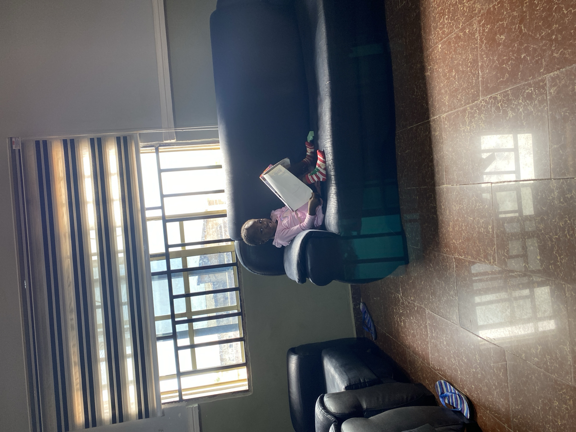 An indoor room, likely in the city of Jos, Nigeria, is depicted with a wide shot encompassing the tiled floor, two dark sofas, and a large window. A young child, appearing to be a girl with dark skin and braided hair, is seated on the larger, three-seater dark blue sofa on the right side of the frame. She is wearing a pink long-sleeved top with ruffled shoulders and striped socks (red, white, and green). The child holds a large white object, possibly a book or container, in front of her, facing slightly towards the left with a subtle smile.
To the left of the frame, a single-seater dark blue armchair is partially visible, and another matching armchair is partially visible in the bottom-left corner. On the reddish-brown tiled floor, which has a mottled, lighter pattern, a pair of blue and white striped flip-flops rests near the left armchair. Another pair of blue flip-flops is visible further back. The floor surface exhibits reflections of the window, indicating a polished finish.
A large window with dark horizontal security bars is prominent in the upper left and center background, allowing abundant natural daylight to illuminate the room. Above the window, a zebra-style day-night blind with alternating translucent and dark grey/blue horizontal stripes is partially drawn. A pull cord for the blind hangs on the right side of the window. The room's walls are painted a light olive-green or grey-green, with a white decorative trim running horizontally below the window level on the right wall. A small, light green object rests on the sofa next to the child's right leg. No other individuals are present or interacting with the child.