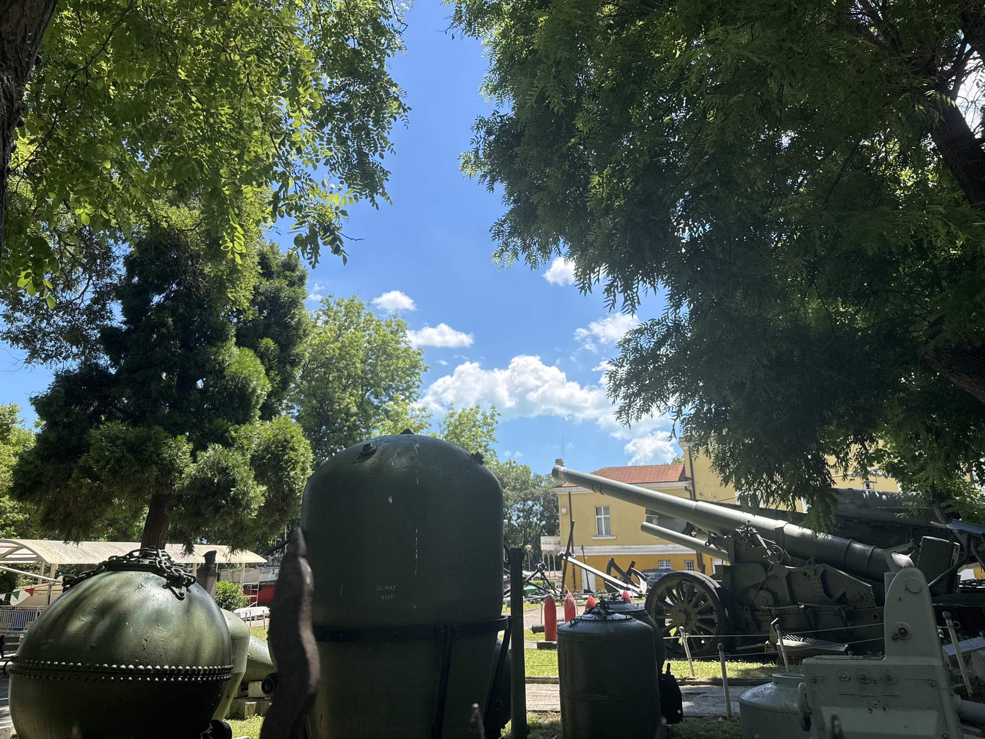 This image depicts an outdoor display of historical military artifacts, likely at a museum or memorial site in Varna, Bulgaria. The foreground is dominated by large, dark green cylindrical objects, one of which has a chain secured to its top. Behind these, a large artillery piece is prominently displayed, featuring a long barrel and heavy wheels. Further in the background, a yellow building with a red-tiled roof stands, suggesting it might be part of the museum's infrastructure. Lush green trees frame the scene, their leaves partially obscuring the bright blue sky dotted with fluffy white clouds. The overall impression is that of a sunny day, with the military equipment placed on a grassy area. There are no people visible in the image.