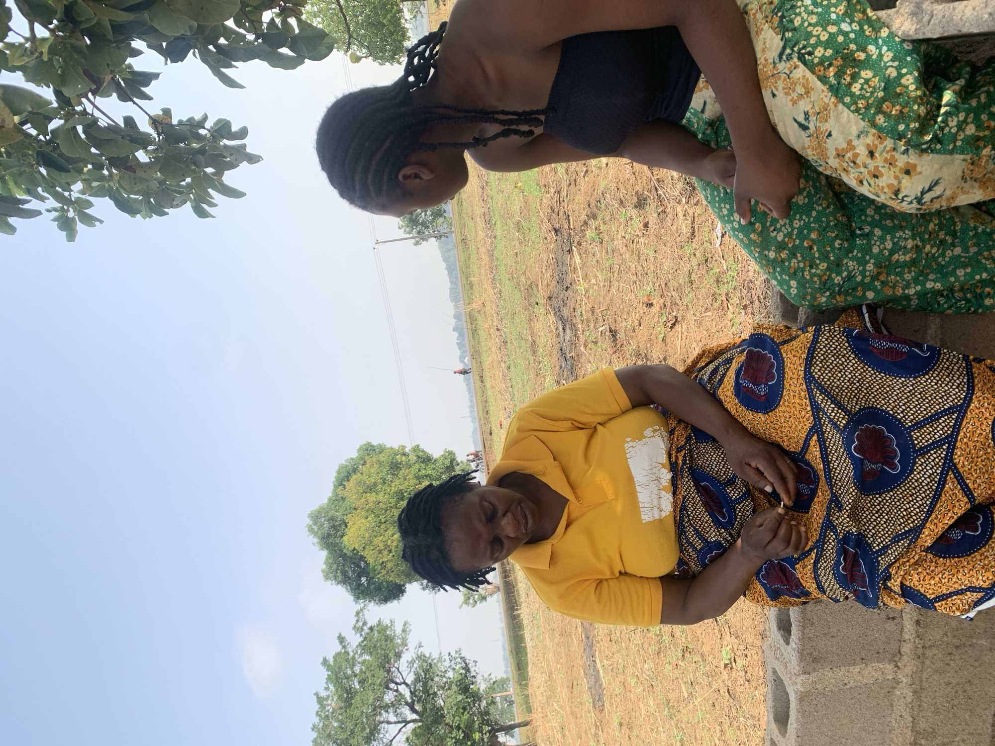 This image, taken in Agban, Nigeria, shows two women interacting in a bright, outdoor setting under a clear sky. The scene suggests a casual conversation or shared moment, likely during the daytime.
In the foreground, an older woman with dark, short braided hair is seated or crouching. She wears a vibrant yellow collared short-sleeved shirt and a colorful skirt adorned with a bold pattern of yellow, blue, and reddish-brown motifs. She looks forward with a gentle, soft smile, and her hands are clasped in her lap, appearing to hold a small, light-colored object or thread. Faded white text or a design is partially visible on the front of her yellow shirt.
Positioned slightly higher and facing the older woman is a younger woman with long, dark braided hair. She wears a black strapless top and a green and cream-colored skirt with a floral pattern. Her profile is visible as she looks down towards the older woman.
The background depicts a rural or semi-rural landscape. The ground is dry, brown earth with sparse vegetation. In the mid-ground and distant background, there are various trees and open fields. Utility poles and lines are faintly visible stretching across the horizon, and a very small, distant figure can be seen in the field. Overhanging green foliage from a tree is visible in the upper left, providing some shade. Simple concrete blocks are situated in the lower right, possibly forming part of a low structure where the women are seated. The weather is sunny with a bright, pale blue sky, indicating a clear day. The overall mood is calm and informal.