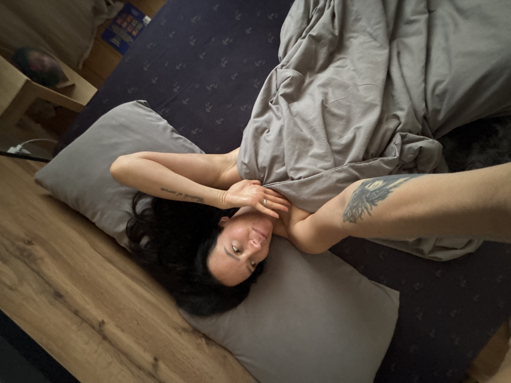 A woman with dark hair lies on a bed with gray pillows and a gray duvet. The bed has a dark blue patterned sheet. The woman is partially under the duvet, revealing her upper body and arms, which have tattoos. She is wearing a ring on her left hand. The room includes a wooden floor and furniture. The location is Chaikovsky, Russia.