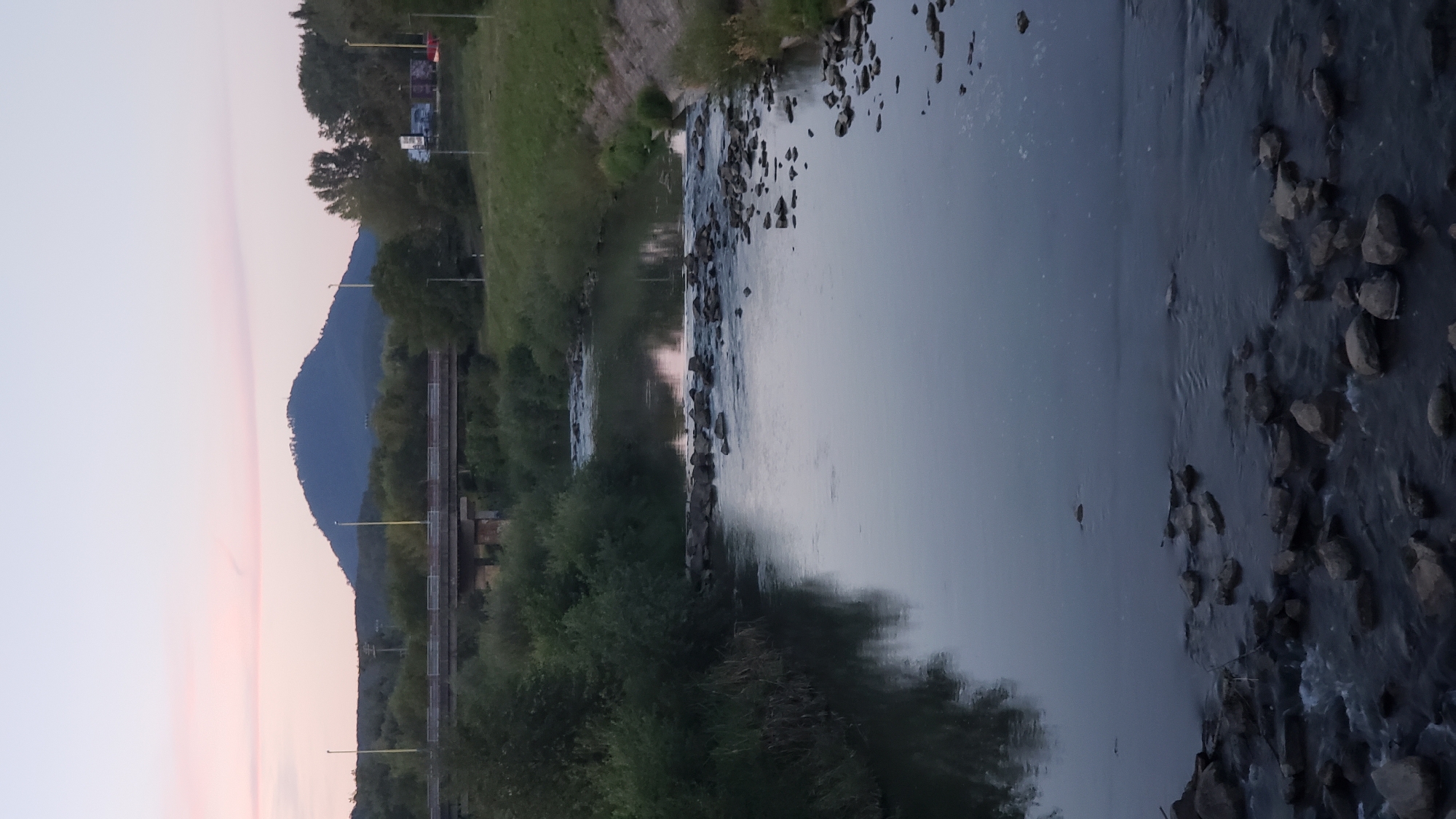 The image depicts a calm river flowing through a natural landscape, possibly at dusk. In the background, a large, dark, conical hill stands against a pale pink sky. A bridge spans the river in the mid-ground, with trees and greenery lining the banks. The riverbed is rocky, with some stones visible above the water's surface, creating small ripples. The water reflects the muted colors of the sky and surrounding vegetation. The scene suggests a peaceful, somewhat rural setting within or near the city of Prešov, Slovakia, given the characteristic conical shape of hills often found in the region.