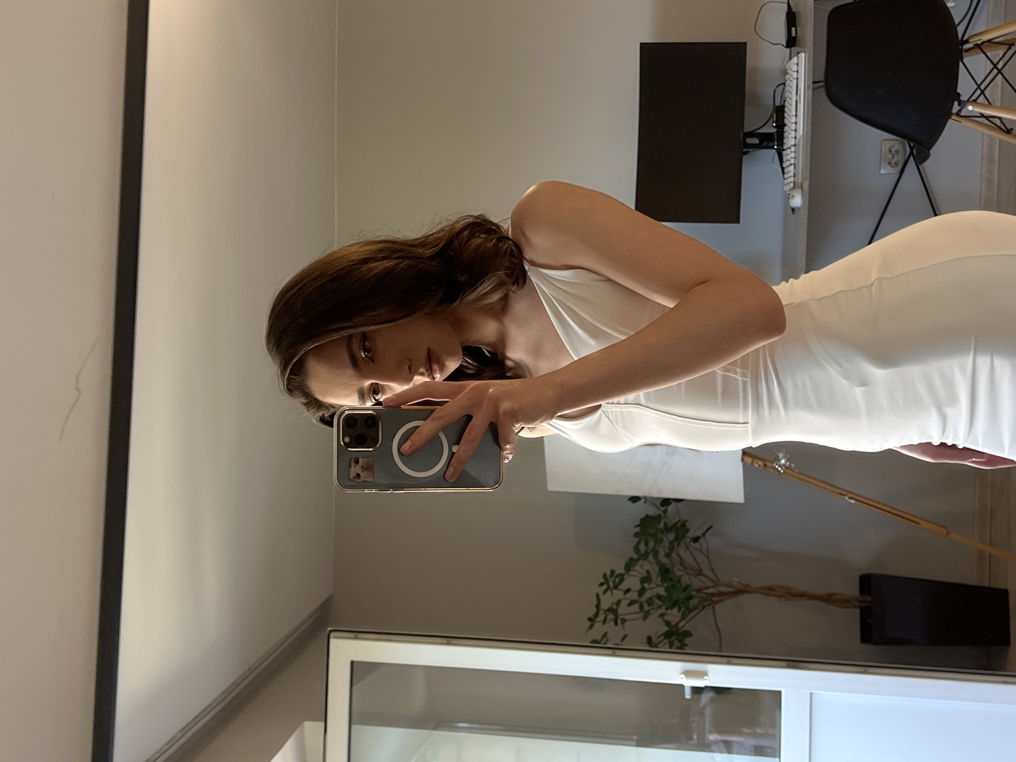 In Ternopil, Ukraine, a young woman with brown hair takes a selfie in a mirror. She is wearing a white, form-fitting dress with thin straps. Her phone, which has a case with a cat sticker and a circular design, is held up to capture the reflection.
The environment appears to be an indoor space, possibly a room in a home or apartment. To her left, a large white door or closet is visible, along with a potted plant. In the background, a desk with a computer monitor, keyboard, and mouse is set up, along with a black chair. A lamp with a wooden stand is also present. The walls are a neutral color.