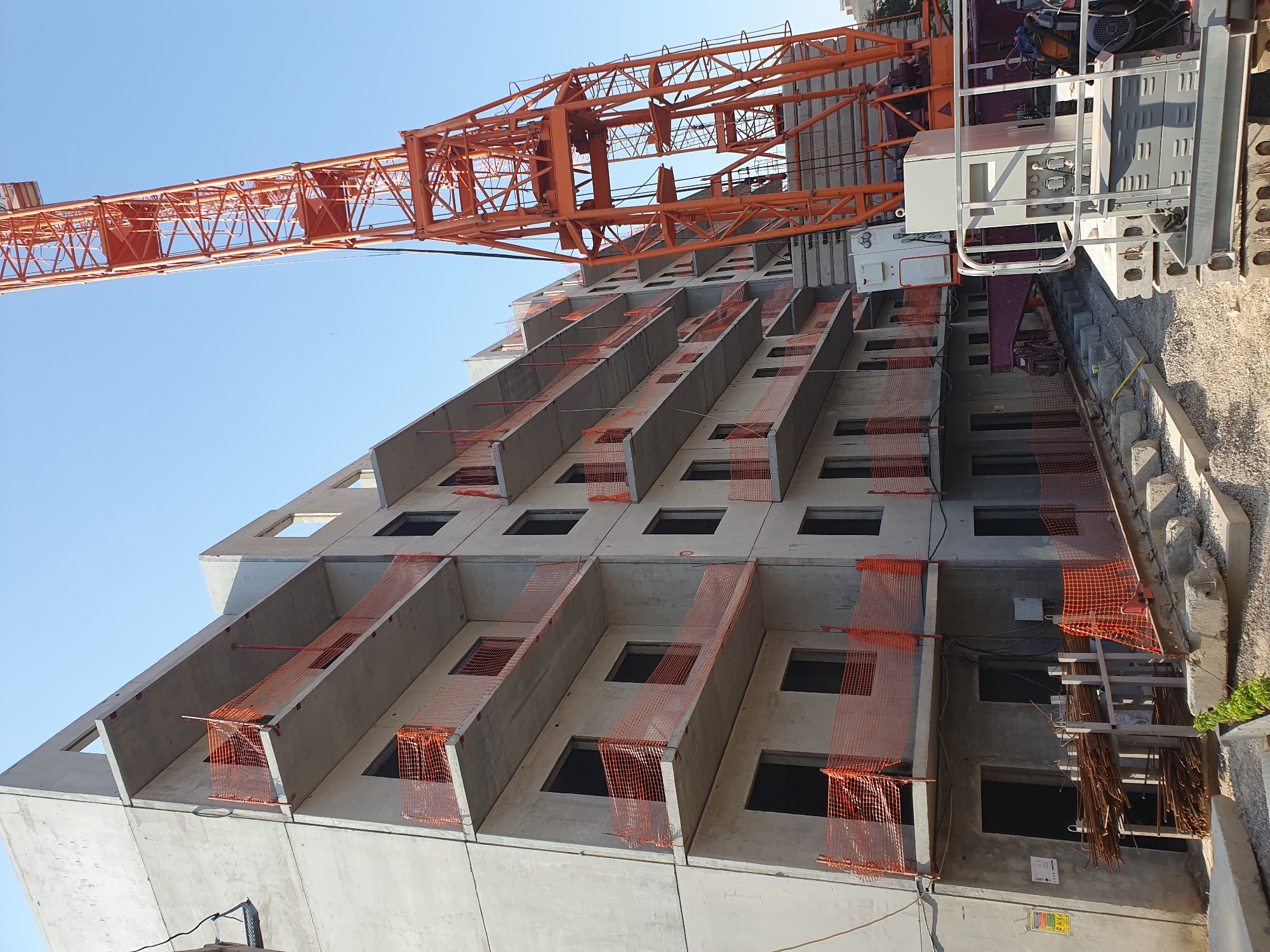 The image captures a construction site in Tashkent, Uzbekistan, focusing on a multi-story concrete building under construction and a large orange tower crane. The building appears to be in an advanced stage, with concrete balconies on multiple floors protected by orange safety netting. The windows and doorways are empty openings. In the foreground, there are stacks of rebar, construction materials, and concrete blocks. The tower crane, a dominant feature, is positioned to the right of the building, with its boom extending upwards. The sky is clear and blue, indicating a sunny day. The overall scene is one of active construction and development in an urban setting.