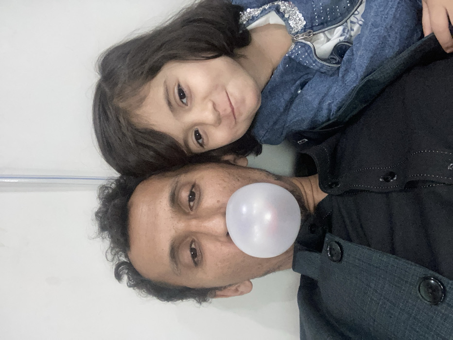 This indoor photograph depicts an adult male and a young female child in Quetta City Tehsil, Pakistan. The adult male, with dark, curly hair and a medium complexion, is situated on the left, wearing a dark-colored, buttoned garment. He is actively blowing a spherical, translucent bubblegum bubble from his mouth. The female child, positioned on the right and leaning against the adult male's left shoulder, has dark, shoulder-length hair and a lighter complexion, smiling towards the camera. She is dressed in a blue denim-style jacket adorned with metallic embellishments on the collar and shoulders, worn over a white patterned shirt. The background consists of a plain, light-colored wall, with a faint vertical line visible towards the upper right.