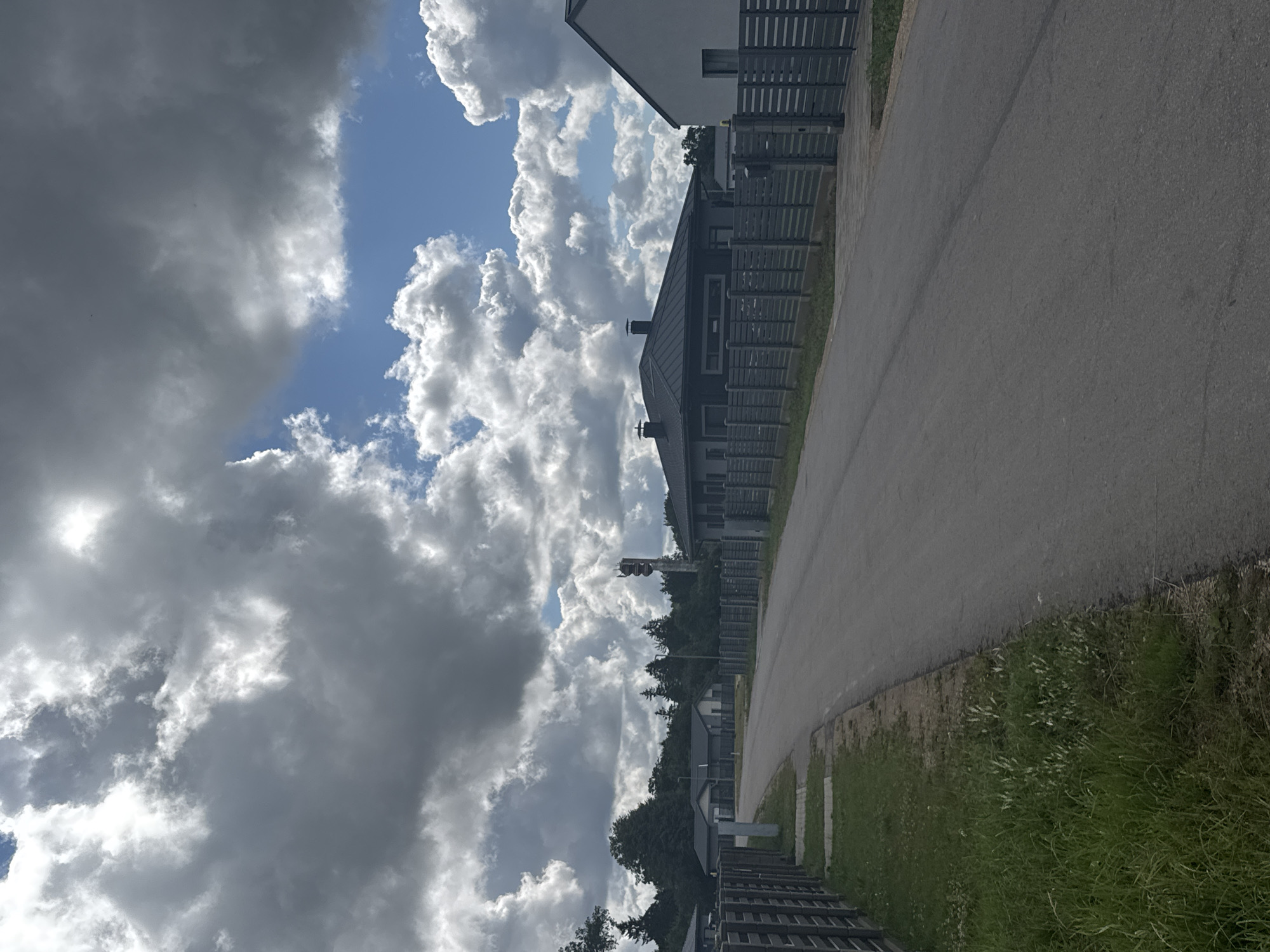 A paved road in Carnikava, Latvia, stretches into the distance, flanked by modern, dark-roofed houses and gray wooden fences. The sky overhead is a dramatic mix of dark, heavy clouds and patches of bright blue sky, with fluffy white cumulus clouds scattered throughout. A tall, slender telecommunications tower with red and white lights is visible in the distance, positioned behind a line of trees. The houses appear to be single-family residences with tidy lawns and driveways. The overall scene suggests a suburban or semi-rural setting on a partly cloudy day.