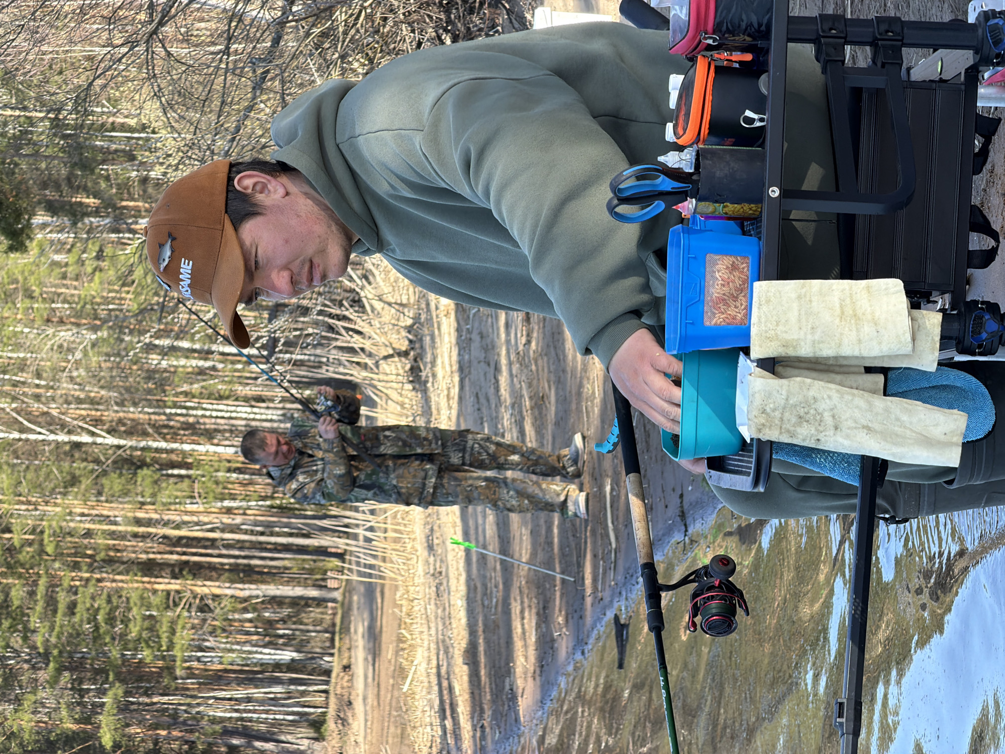 The image depicts two men engaged in fishing activity, likely in an outdoor setting near a body of water, which is consistent with being near Chaykovsky, Russia, a city known for its proximity to the Kama River.
The foreground features a man with a brown cap and a green hoodie. He is leaning towards a fishing station consisting of various containers, including a blue container with what appears to be bait. He is reaching for a teal container.
A fishing rod is set up in front of him.
In the background stands another man wearing camouflage clothing, also fishing with a rod.
The surrounding environment shows signs of early spring or late autumn with bare trees and vegetation, suggesting a riverside location. A green arrow is on the ground near the background man.