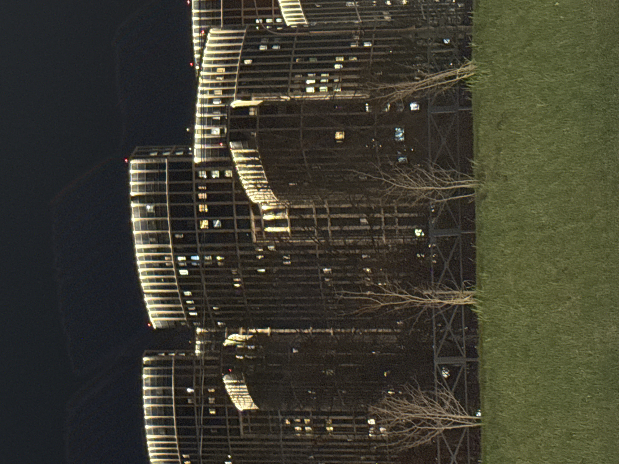 The image shows a night scene of buildings with lights in Khimki, Russia. The structures appear to be tall, possibly residential, with numerous illuminated windows. Adjacent to the buildings is a strip of green space, likely grass, and leafless trees. The background consists of a dark, undefined area, suggesting it is nighttime.