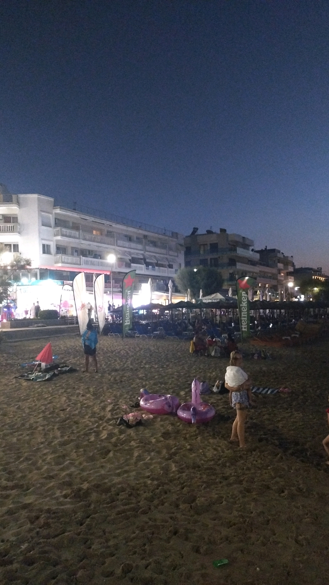 In Perea, Greece, the scene is a beach at dusk, with the dark sky providing a backdrop for illuminated buildings and beach amenities. Several buildings line the background, appearing to be hotels or apartments with balconies and lights. In the middle ground, a row of beach umbrellas and chairs are set up, suggesting a commercial establishment. Several flags are visible, including ones with the "Heineken" logo. On the beach itself, there are scattered beach towels and inflatable toys, including two pink flamingo-shaped floats. A few people are visible on the beach: a person in a blue top and dark shorts stands near the middle of the frame, facing away from the camera; a woman in a patterned dress is holding a child; and other individuals are seen in the distance, seated or walking. The sand is the primary ground cover, showing footprints and ripples. The overall atmosphere is relaxed, possibly indicating the end of a day at the beach or the beginning of an evening gathering.