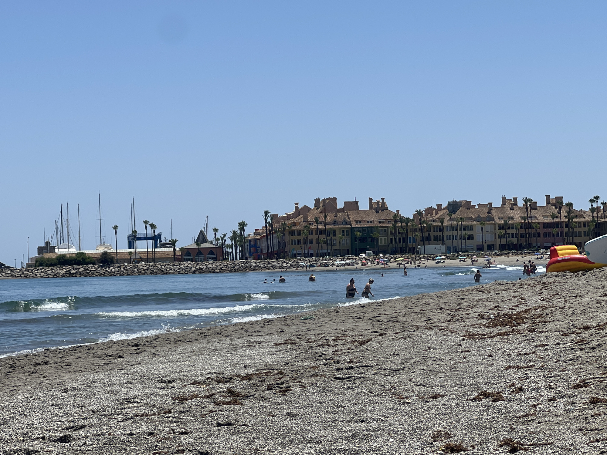 The image shows a beach scene in San Roque, Spain, with the ocean in the foreground and buildings and a marina in the background. The beach is covered in pebbles and seaweed. Several people are in the water, swimming and wading. Some people are on the beach, sunbathing and relaxing. An inflatable water toy, red and yellow, is partially visible on the right side of the image. The buildings in the background appear to be apartments or hotels, with terracotta roofs and palm trees in front of them. The marina features several sailboats and other vessels. The sky is clear and blue, indicating a sunny day.