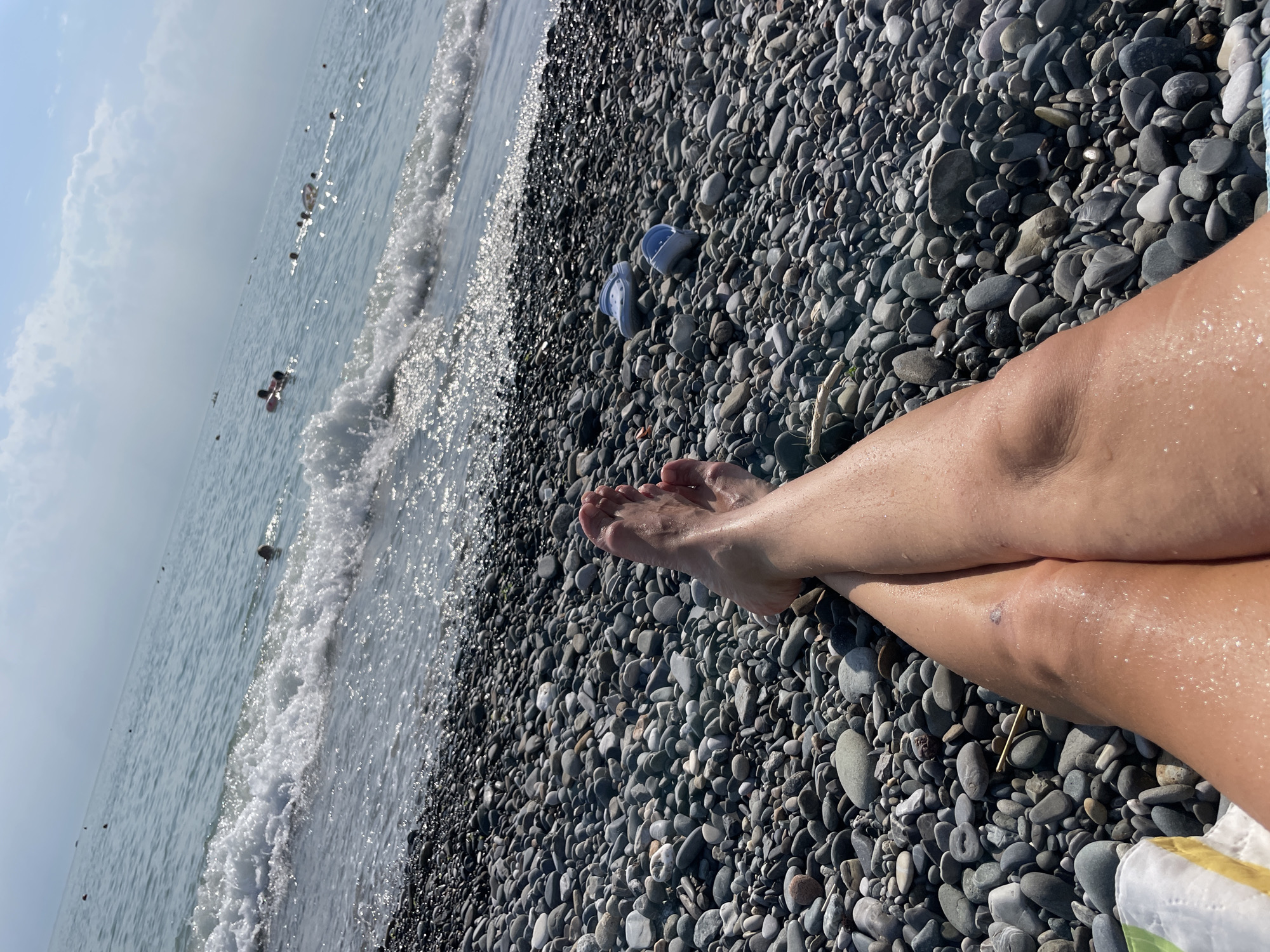 A first-person perspective shot of a person's legs and feet resting on a pebble beach. The waves of the Black Sea are in the background with several people swimming. Two blue flip-flops are visible on the pebbles to the right of the frame.