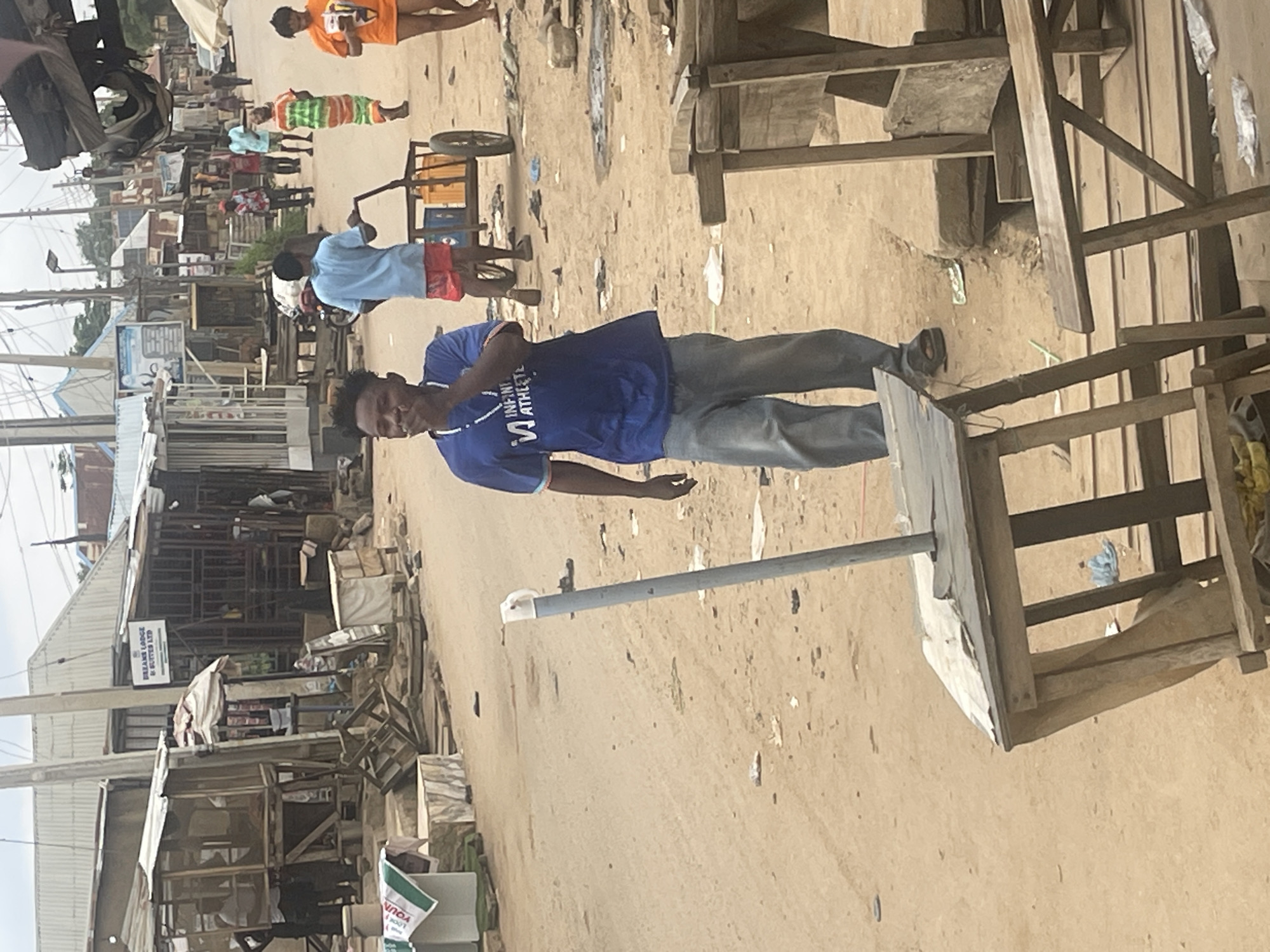 The image depicts a street scene in what appears to be a market area in Abuja, Nigeria. The foreground is dominated by a young Black man wearing a blue t-shirt with white text that reads "INFINITE ATHLETE" and grey pants. He is standing and looking towards the viewer, with his right hand slightly raised. Behind him, a grey metal pole with white fixtures runs diagonally across the frame.
The background reveals a bustling environment with numerous stalls and structures made of wood and corrugated metal. Several other people are visible in the distance, some standing, others walking. One person in an orange t-shirt is seen towards the top of the frame, and another in a blue shirt and red shorts is further back. A cart with wheels and a yellow container is also present. The ground is dusty and uneven, indicative of a street or open market.
Several signs are visible on the buildings. One prominent sign on a white facade reads "DREAMS LODGE & SUITES LTD". Another sign, partially visible at the bottom right, has some green text and the word "LOOK". The scene suggests a typical daytime activity in a commercial or residential area. The lighting indicates it is daytime, and the weather appears to be fair. The overall impression is one of a lively, everyday street scene in an urban Nigerian setting.
