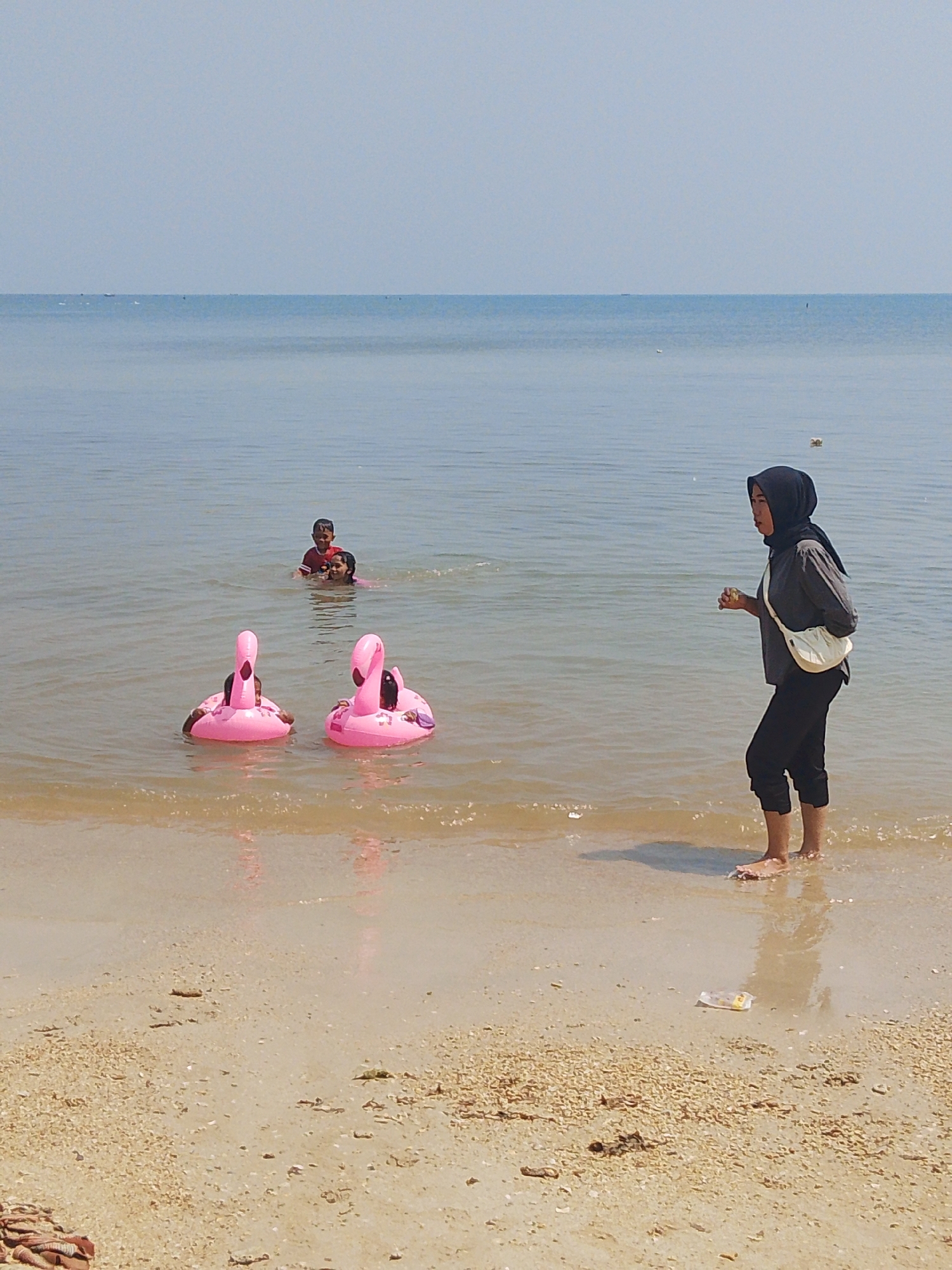 In the city of Pejagan, Indonesia, a woman wearing a dark hijab, gray shirt, and black pants stands ankle-deep in the calm, shallow waters of a beach. She holds a small, light-colored object in her right hand and has a cream-colored bag slung across her body. To her left, two pink flamingo-shaped inflatable floats are in the water, each with a child inside. Further out in the water, two children are visible, one appearing to be an adult in the water with a child on their shoulders. The sky is clear and pale blue, meeting the horizon of the vast, tranquil sea. The beach in the foreground is covered in light-colored sand and small debris.