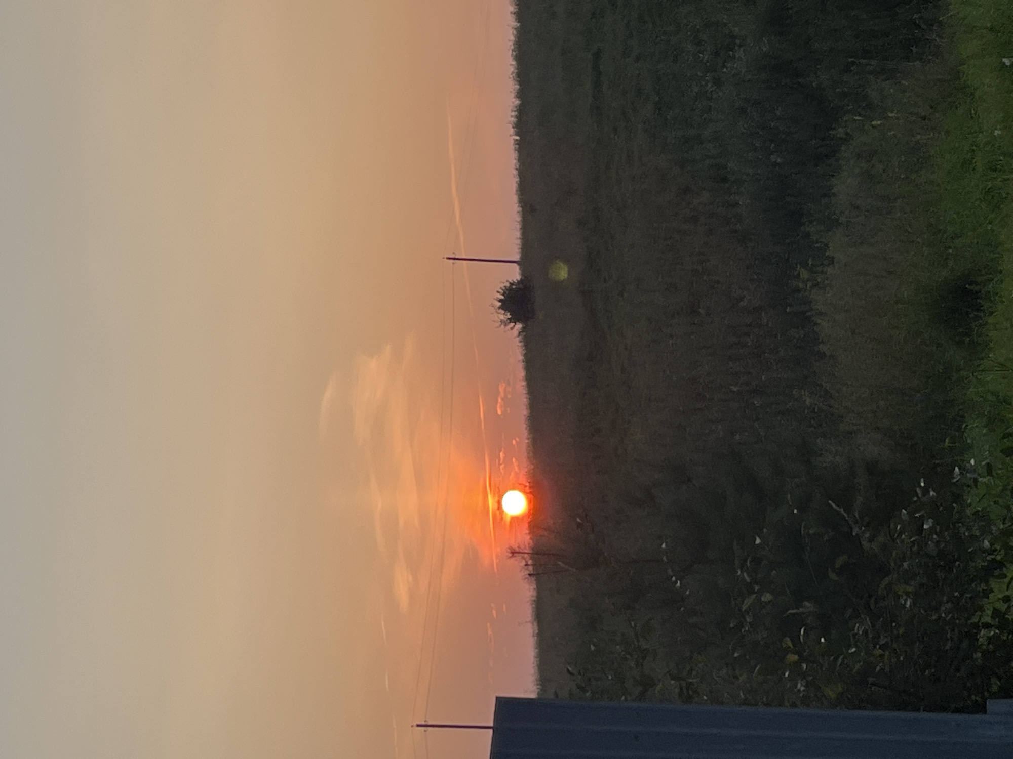 The image depicts a rural landscape with a sunset in the background. The sun, a bright orange orb, is low in the sky, casting a warm glow over the scene. Wispy clouds are visible, tinged with the colors of the sunset. A grassy field stretches across the foreground, dotted with vegetation. Two utility poles are present, one on the left and another in the distance. A dark, blocky structure is partially visible on the far left edge of the frame. The overall impression is one of tranquility and natural beauty.