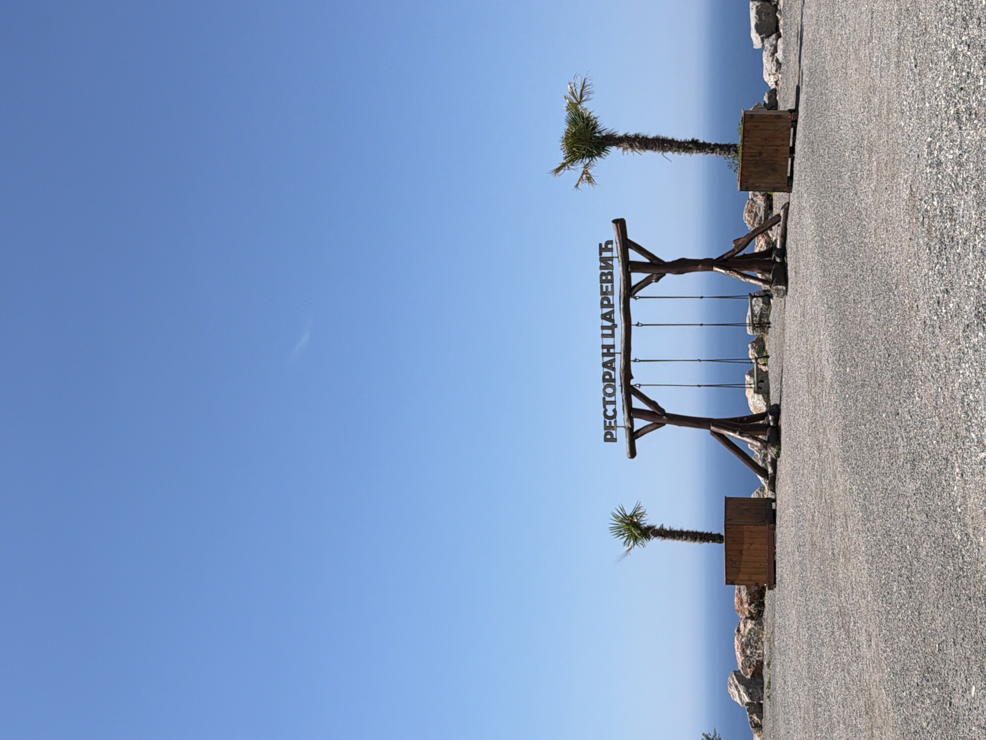 The image depicts a clear blue sky over a coastal landscape. In the foreground, a gravelly surface is visible, leading towards a wooden swing set with the Cyrillic inscription "РЕСТОРАН ЦАРЕВИЂ" (Restoran Carevic) above it. Two palm trees, one smaller and in a wooden planter on the left, and a taller one in a planter on the right, flank the swing set. Beyond the swing set, a rocky embankment drops off towards the deep blue sea. The horizon line is defined by the meeting of the sea and the sky. The location is identified as Kovačko Polje, Montenegro. There are no people or animals visible in the scene.
