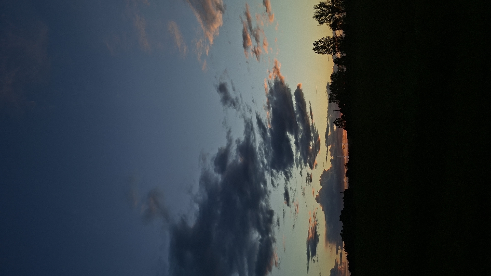 The image shows a vertical, slightly rotated view of a dusk or dawn scene. A dark, mostly obscured landscape occupies the right side, suggesting either dense vegetation or complete darkness. A line of trees is silhouetted against the lighter sky. The sky transitions from a deep blue on the left to lighter blues and oranges towards the right. There are prominent cloud formations throughout, some dark and stormy-looking, others tinted with orange and pink hues from the sun. Power lines are barely visible along the horizon. The location is Забалоцкі сельскі Савет, Belarus.