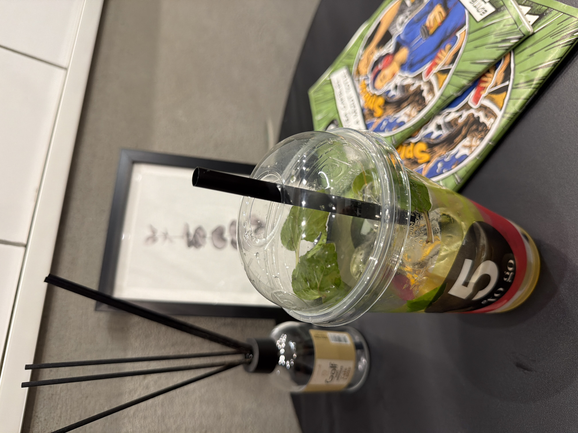 A clear plastic cup containing a beverage with mint leaves and ice is positioned on a dark gray table. A black straw extends from the cup's lid. To the left of the cup is a glass bottle with black reeds, commonly used as a room diffuser. Behind the diffuser, a framed picture hangs on a gray wall. To the right of the cup, two magazines with colorful covers are stacked. The environment appears to be indoors, possibly a cafe or home, with a white tiled ceiling visible at the top of the frame. The lighting suggests an indoor setting.