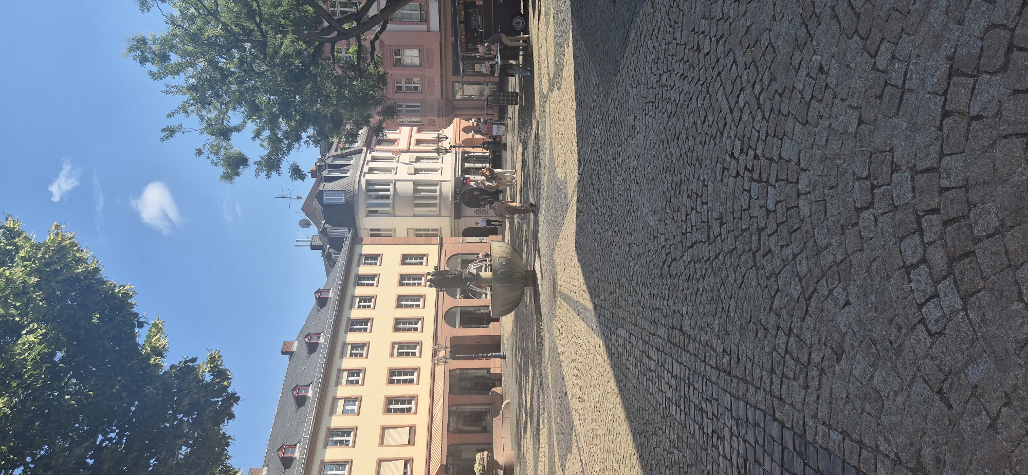 This is a vertical, eye-level shot of the Kirschgarten square in Mainz, Germany, taken on a sunny day. The square is paved with cobblestones, which are laid out in concentric semi-circles, with a prominent shadow cast across the foreground from an unseen object to the right. In the middle ground, a large, ornate stone fountain with sculpted figures is active, with water flowing. Behind the fountain, a row of multi-story buildings forms the backdrop. The buildings have a light yellow facade with dark red accents around the windows and on the ground floor. One building to the right of the yellow one is pale pink, and another further right is a soft white. Several people are visible in the square, some walking and others standing, interacting with each other or observing the fountain. A few trees are present, one large one to the right with lush green leaves, and branches of another tree are visible in the upper left corner, framing the bright blue sky with a few wispy clouds. The overall atmosphere appears to be one of a pleasant, public urban space.