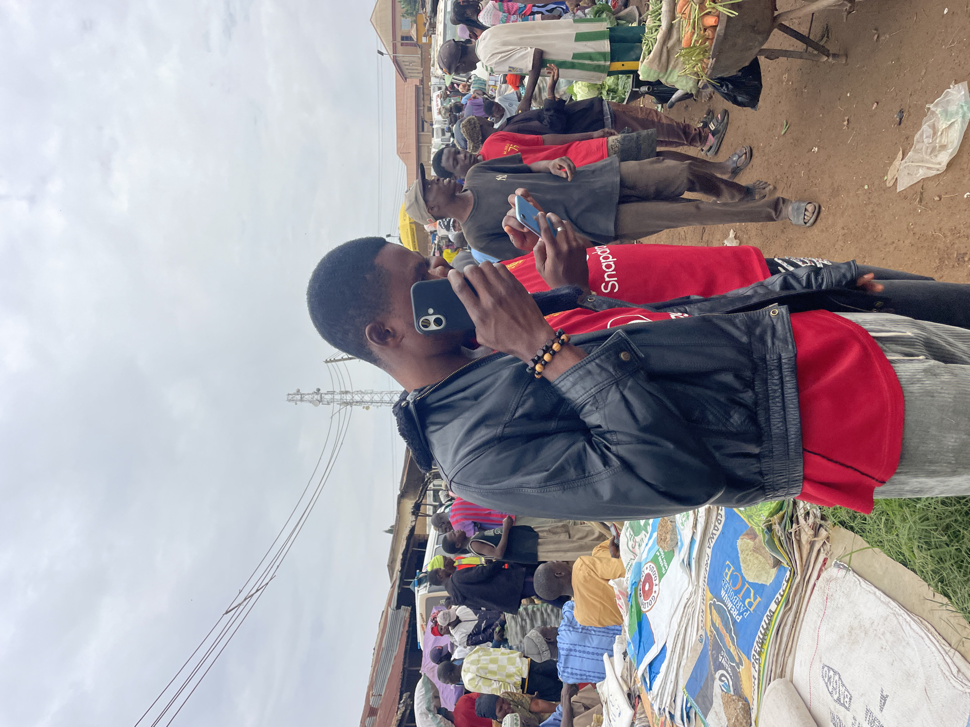 The image depicts an outdoor market scene in Gura Top, Nigeria, under an overcast sky. The foreground is dominated by a young Black man in a black leather jacket and a red t-shirt, holding up a smartphone and appearing to take a picture or video. To his left, another young Black man in a red t-shirt with white lettering, possibly "Snapdragon," is also holding a smartphone.
The background reveals a bustling marketplace with numerous individuals, stalls, and goods. Various people are seen dressed in casual clothing, some wearing traditional headwear. Stalls display a variety of products, including bags of rice and fresh produce like carrots. In the middle ground, a few men are standing and conversing, while others are seen walking through the market. In the distance, buildings and a white van are visible, suggesting an urban setting. The ground is dirt, indicating a typical market terrain. The overall atmosphere suggests a lively and active commercial environment.