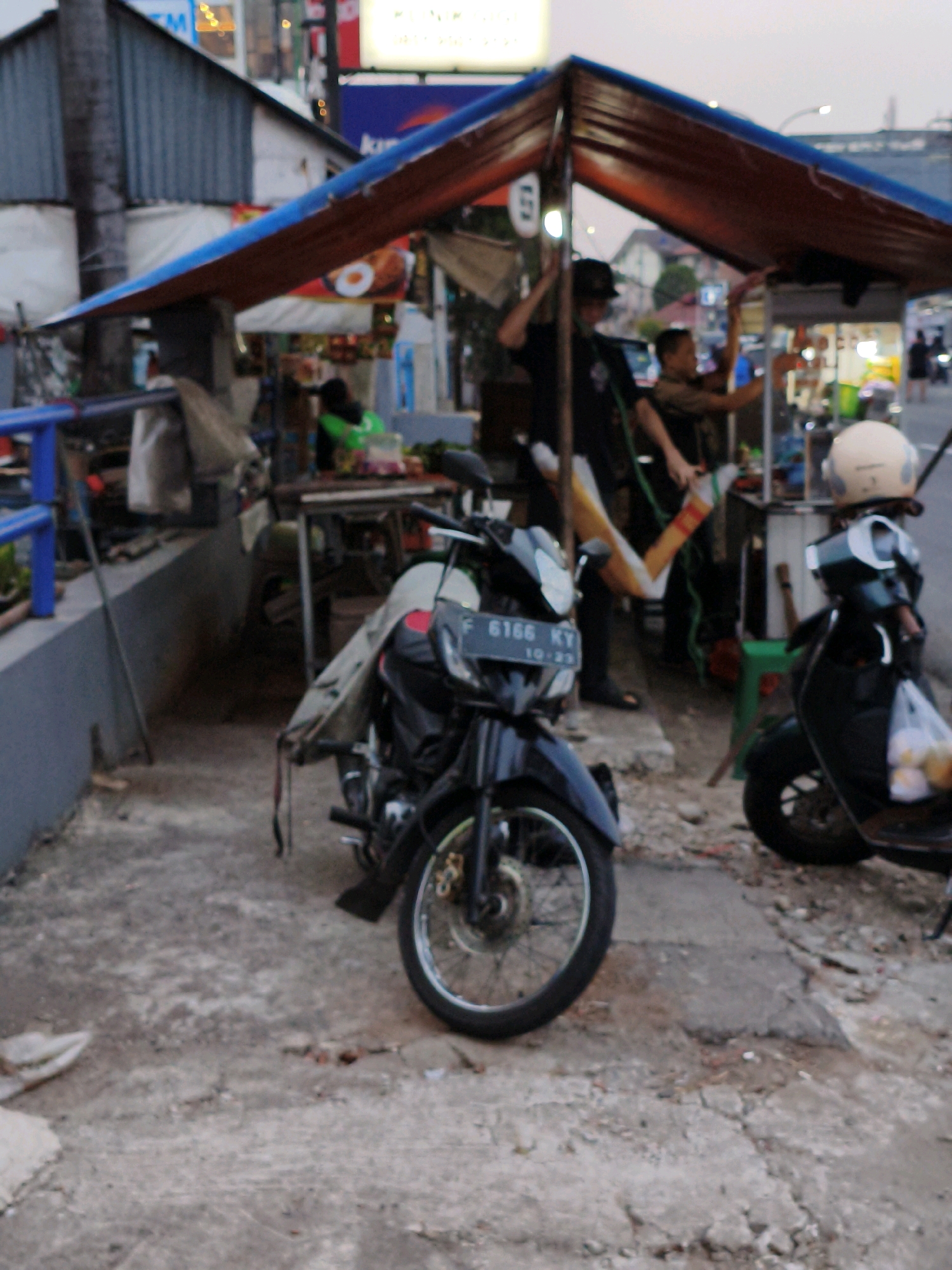The image depicts a street scene in Cibinong, Indonesia, likely during twilight. In the foreground, two parked motorcycles are visible. The motorcycle on the left is black and white with a license plate reading "F 6166 KY 10-23". The motorcycle on the right is also black with a light-colored helmet resting on its seat. Behind the motorcycles, a food stall with a blue and red awning is set up, where two individuals are interacting. One person, wearing a black cap and shirt, appears to be serving or preparing food. Another person in a brown shirt is also present at the stall. The environment includes a paved ground, a building with a corrugated metal roof in the background, and street lights visible in the distance. The overall impression is a bustling street-side food vendor operation.