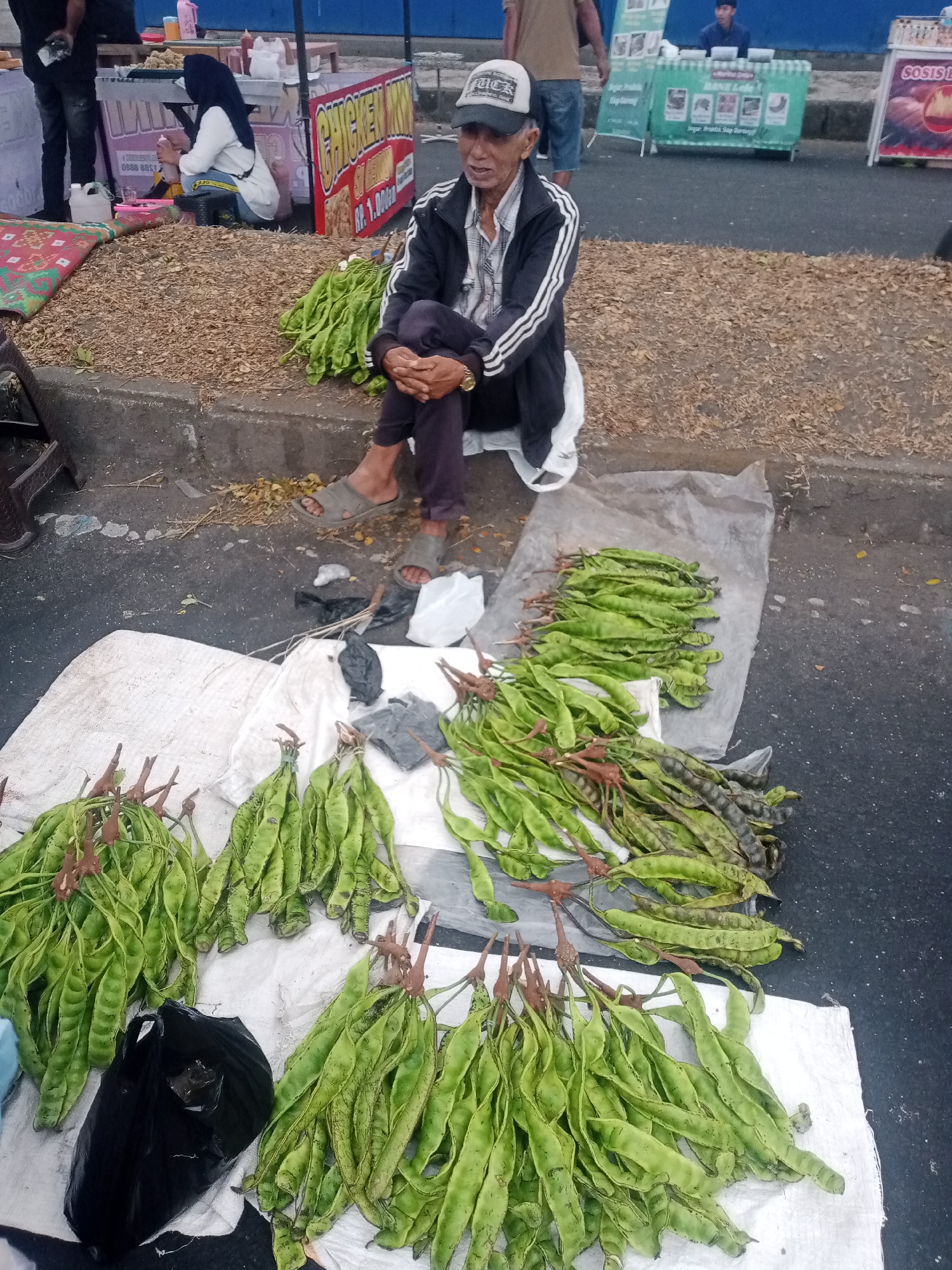 The image depicts a street scene, likely a market, in Tasikmalaya, Indonesia. An elderly man, wearing a cap and a jacket over a plaid shirt, is seated on a curb with his legs crossed. He is surrounded by bundles of green pods, presumably a type of vegetable, arranged on white fabric and a clear plastic sheet on the ground. In the background, there are food stalls with signage indicating items like "CHICKEN MN" and "SOSIS LOKAL". Other individuals are visible in the background, including a woman in a hijab at one stall and a man behind a counter at another. The ground is covered with gravel and debris, suggesting an outdoor, possibly informal, marketplace setting.
