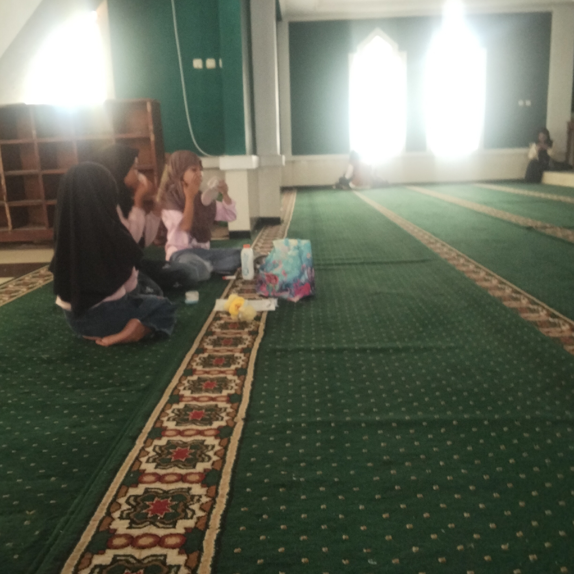 The image depicts an interior scene, likely a mosque, with green carpeted floors and decorative woven borders. In the foreground, two young girls, wearing hijabs, are seated on the floor. One girl is wearing a black hijab and a pink shirt with blue pants, while the other wears a grey hijab and a pink shirt with blue jeans. They appear to be interacting, with one holding a white object. To their right, there is a small white bottle and a colorful patterned bag. In the background, two large windows, possibly stained glass or illuminated from behind, cast a bright light. Shelving units are visible on the left side of the image. The environment suggests a place of worship or community gathering, common in locations like Cisaat, Indonesia.