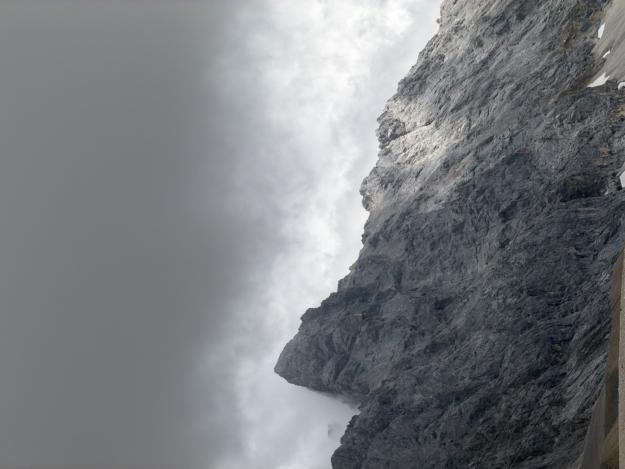 The image depicts a rugged, grey mountain peak shrouded in clouds under a heavy, overcast sky. The mountain's rocky face is textured with deep crevices and sharp edges, with a dramatic slant across the frame. A sliver of sunlight breaks through the clouds, illuminating a portion of the mountain's slope, creating a stark contrast between light and shadow. Patches of snow are visible on the lower slopes. The foreground shows a rocky, barren terrain. This is Zugspitze, the highest peak in Germany, located in Garmisch-Partenkirchen. There are no people or discernible human activity in the image.