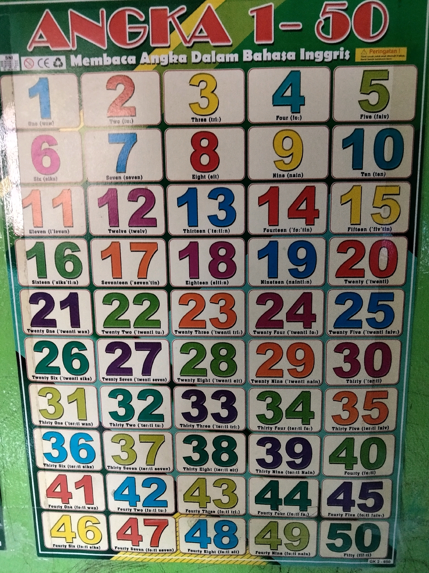 The image displays a colorful educational chart titled "ANGKA 1-50" (Numbers 1-50) written in bold red and yellow letters against a green background. Below the title, in smaller white text, it reads "Membaca Angka Dalam Bahasa Inggris" (Reading Numbers in English). The chart is organized into a grid containing numbers from 1 to 50. Each number is prominently displayed in a large, bold font with varying colors for the numbers themselves (e.g., blue for 1, red for 2, yellow for 3). Below each number, its English pronunciation is provided in smaller text, enclosed in parentheses, such as "(wan)" for 1, "(tu:)" for 2, and "(tri:)" for 3. The chart also contains small logos indicating "SNI" (Indonesian National Standard) and "CE" (Conformité Européenne). A yellow warning triangle with "Peringatan!" (Warning!) is visible in the top right corner, with accompanying Indonesian text. The chart appears to be a physical poster or wall hanging, as indicated by the texture and slight wrinkles. The background of the chart itself is a light cream color, with the grid lines and borders in a darker brown. The overall setting suggests an educational or domestic environment where children might be learning numbers. The location of Rahong, Indonesia, is not explicitly depicted or inferable from the image itself.