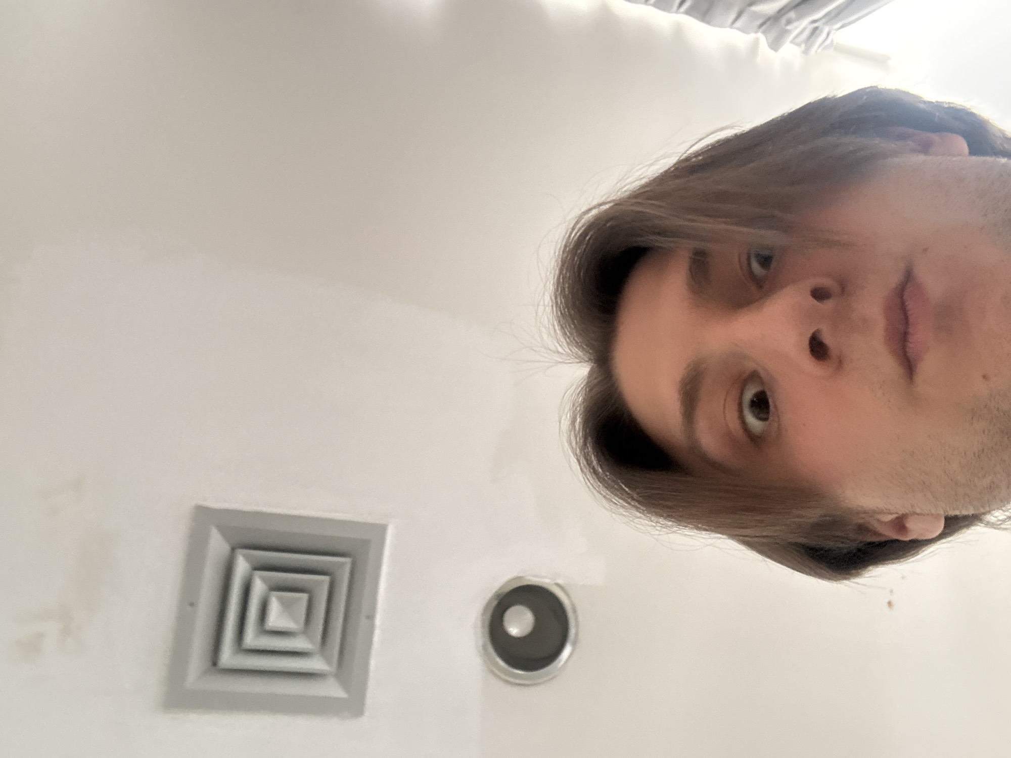 The image displays a close-up, rotated view of a person's face and a white ceiling from a low-angle perspective, suggesting the camera is pointing upwards. The subject, positioned on the right side of the frame, is a person with medium-length brown hair, brown eyes, and light facial stubble. They are looking directly into the camera with a neutral expression.
On the white ceiling, visible on the left side of the frame, there is a square air vent or diffuser with a series of concentric square patterns. Below and to the right of the vent, a smaller, circular object—possibly a recessed light fixture or a sensor—is also present. The ceiling shows some minor, faint smudges or discolorations near the air vent. In the top part of the original image (which becomes the left edge after rotation), a light-colored, possibly striped fabric or bedding material is partially visible, indicating the person may be lying down.
The lighting is soft and even, consistent with an indoor setting. There are no notable details regarding the time of day, weather, or specific location cues within the image itself to confirm the provided context of Milan, Italy. No text is visible.