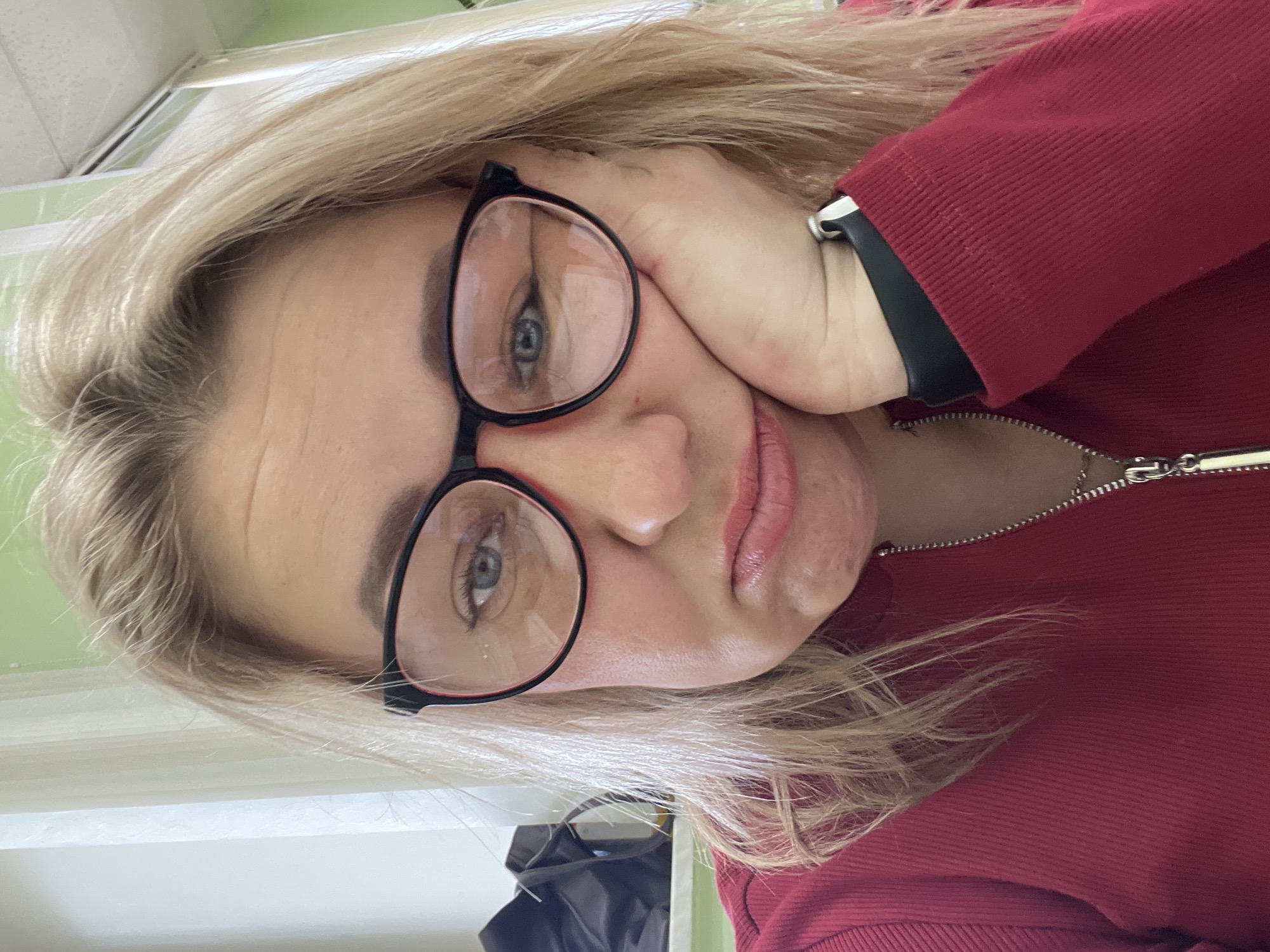 The image features a woman, presumably located in Chaikovsky, Russia, captured in a close-up, slightly upward-angled portrait. She has blonde hair and is wearing black-framed glasses that highlight her blue eyes. Her left hand is raised, supporting her head with fingers resting against her cheek and temple. A black wristwatch is visible on her left wrist. She is dressed in a vibrant red long-sleeved top, featuring a zippered front and a ribbed texture on the sleeves. Her lips are slightly parted, and her expression appears pensive or neutral as she looks towards the camera.
The setting is an indoor environment, with a light-colored ceiling and walls visible in the blurred background. A dark, indistinct object, possibly a piece of furniture or equipment, can be seen in the lower part of the background. The lighting suggests an indoor setting, possibly with artificial illumination or ambient daylight. No specific time of day or weather conditions can be determined from the image. There is no visible text present.
