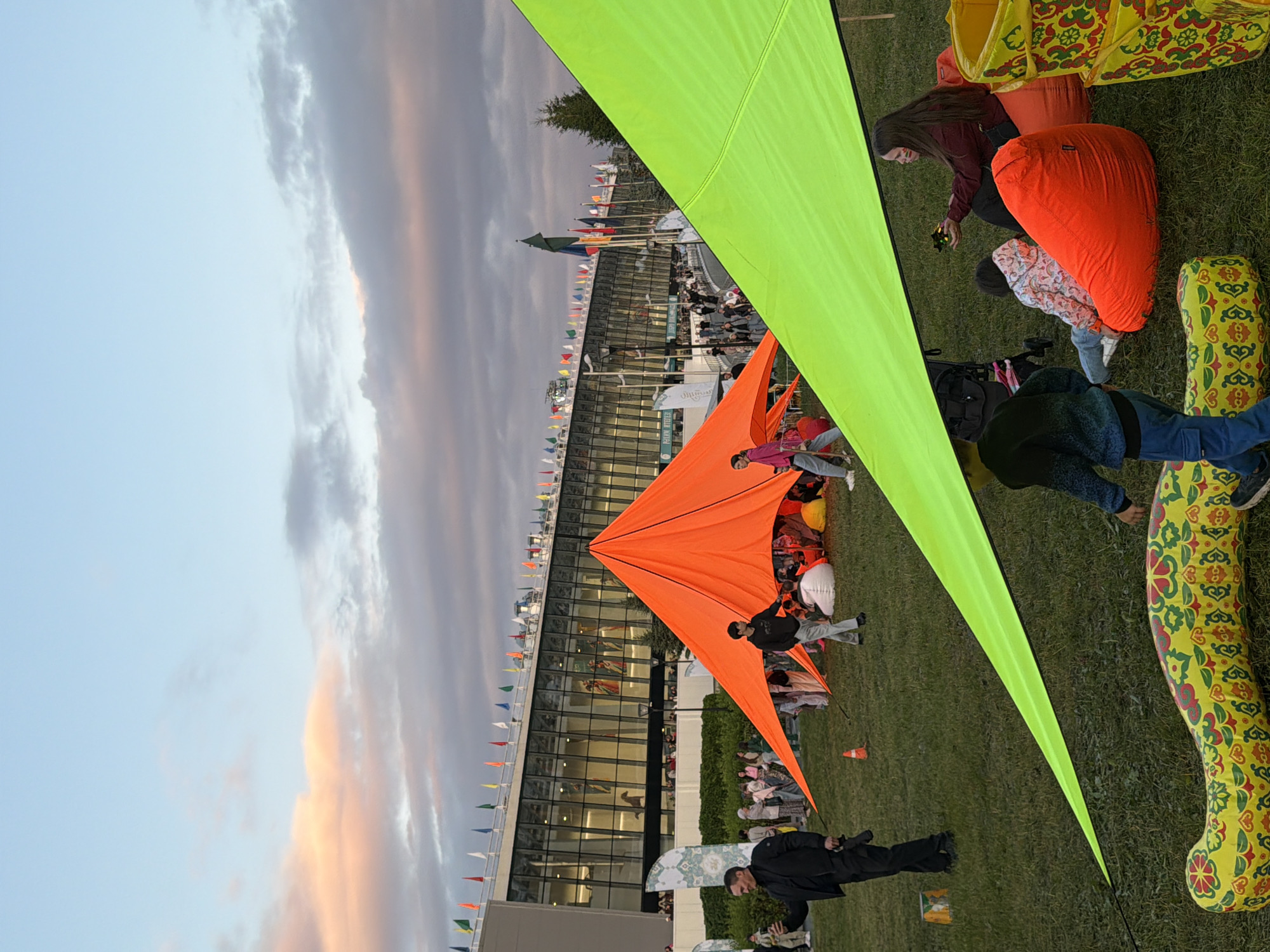 This is a full-shot, eye-level, outdoor photograph taken at dusk in an unknown location. In the foreground, a bright lime green canopy is draped over a grassy area. To the left, an orange, pyramid-shaped tent is set up. Several people are scattered around, some sitting on beanbag chairs under the canopy, others standing near the tents or walking in the background.
In the midground, a man in a black suit stands to the left, holding a camera. Behind him, a crowd of people mills about. To the right of the orange tent, a group of people are gathered, some sitting on beanbags. Further back, a large, modern building with a glass facade stretches across the background, adorned with numerous small flags. The sky above is a soft blue, with wisps of pink and orange clouds, indicating sunset. The ground is covered in green grass, with a yellow and red patterned inflatable toy snake lying horizontally in the lower left corner.
